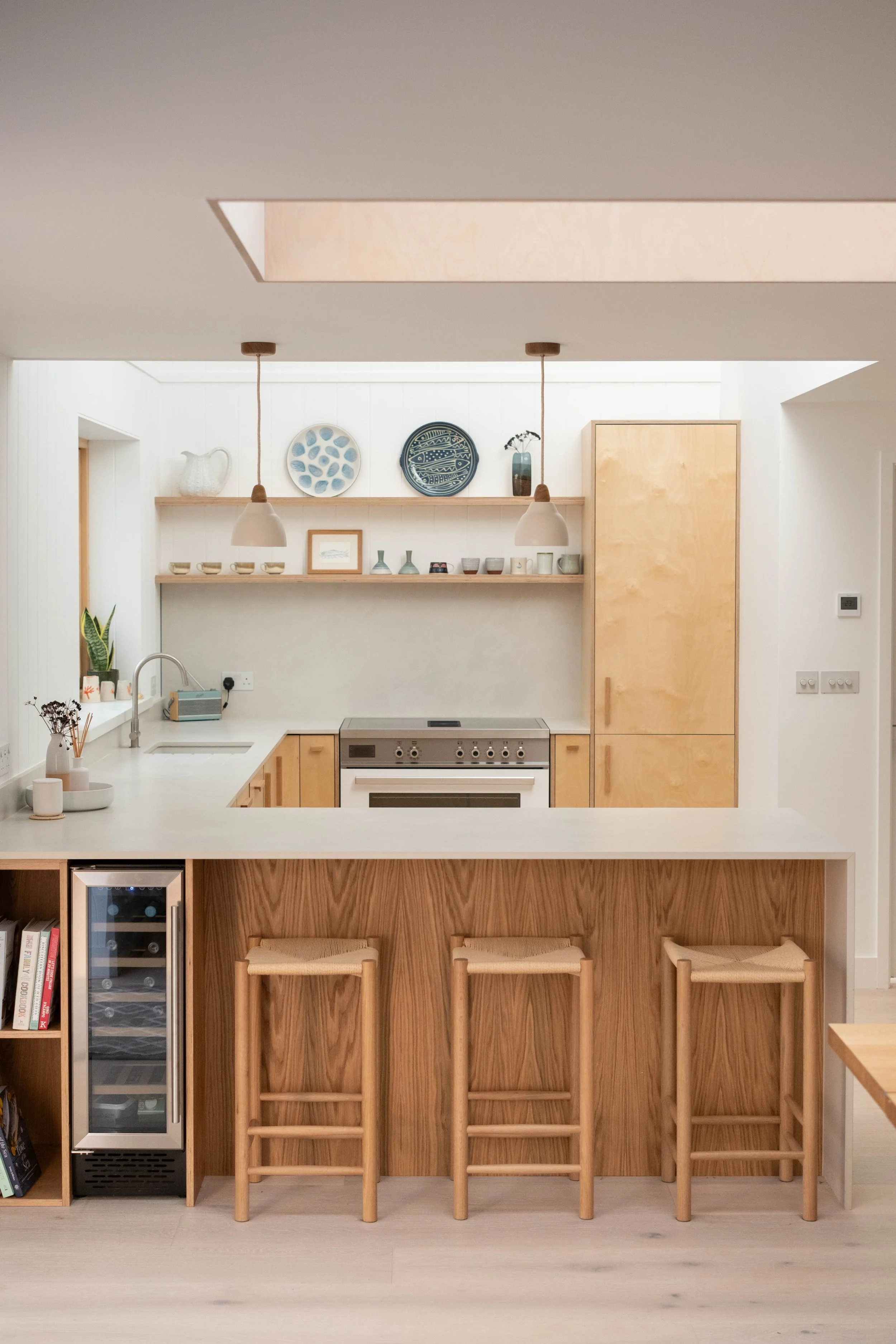 Kitchen with wooden cabinets, white countertop, and a stove. Above, a wooden shelf with decorative plates, vases, and artwork. Two pendant lights hang from the ceiling. A small refrigerator is under the counter, and three wooden stools are at a breakfast bar.
