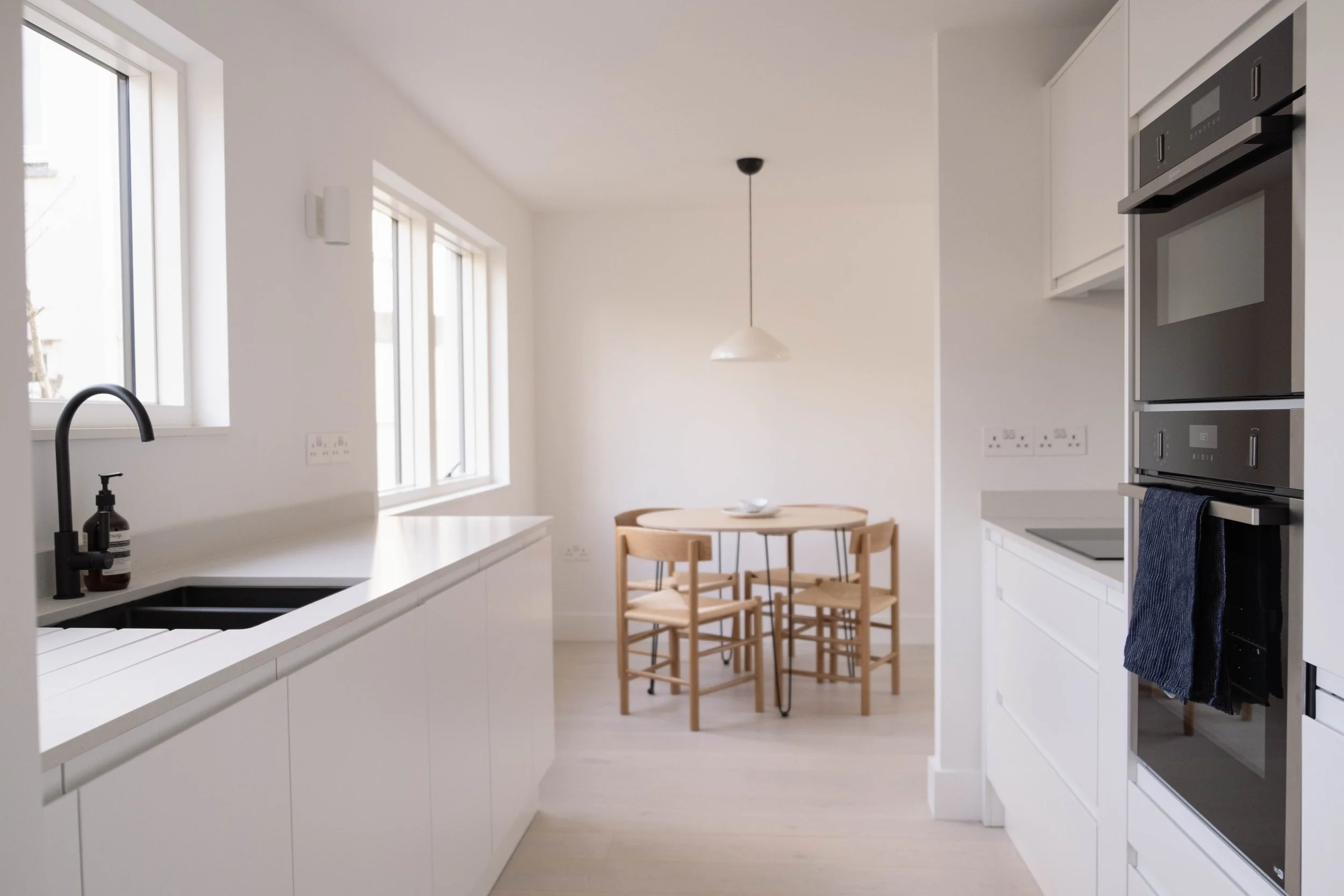 Interior view of a modern kitchen with white cabinets, black sink, and countertop, wooden dining table with four chairs, and built-in oven and microwave, with large windows letting in natural light.