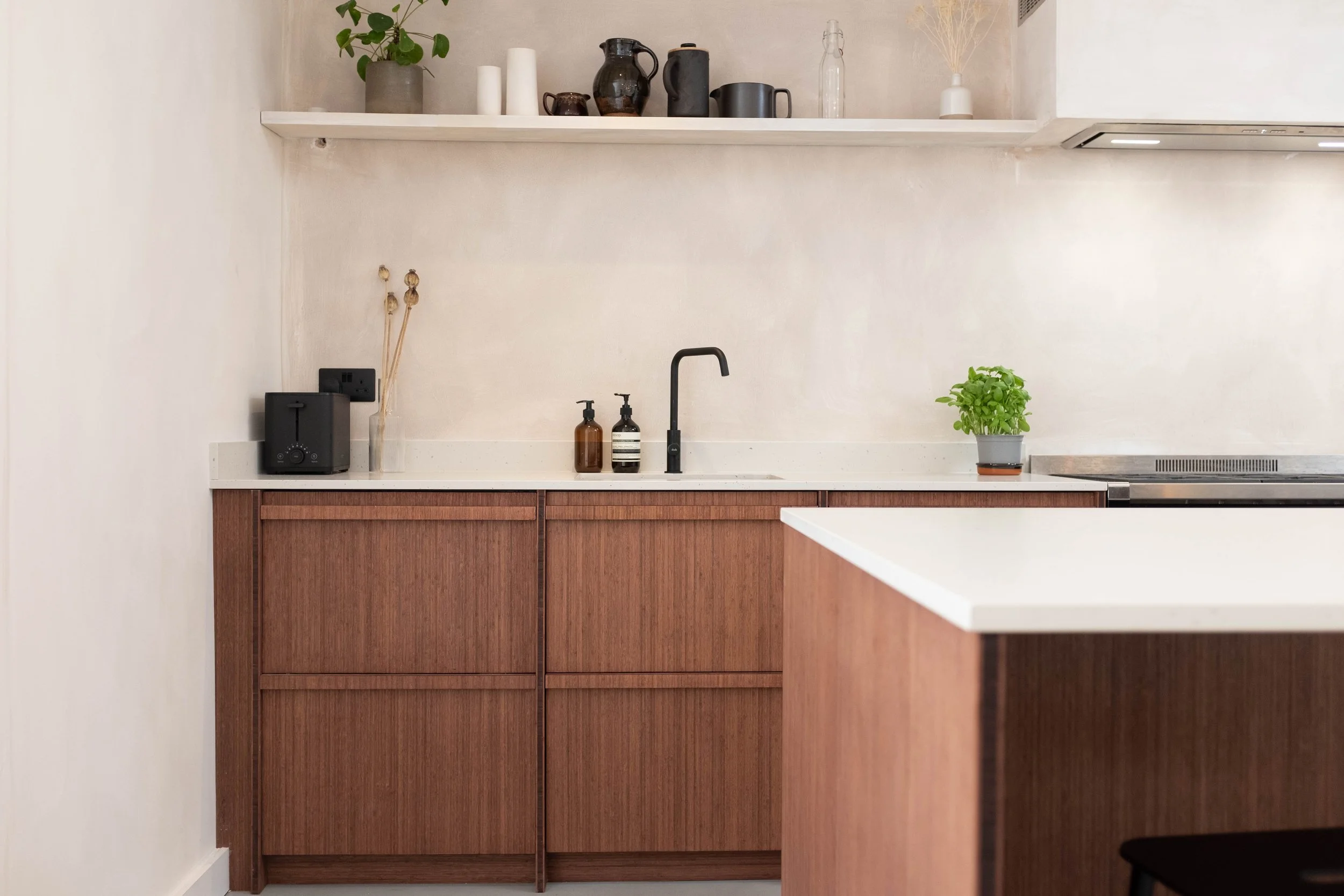 A modern kitchen counter with a black faucet, two soap dispensers, a green potted herb, and a wooden cabinet with a white countertop. There are decorative vases and jars on a white shelf above, and a small black appliance on the left.
