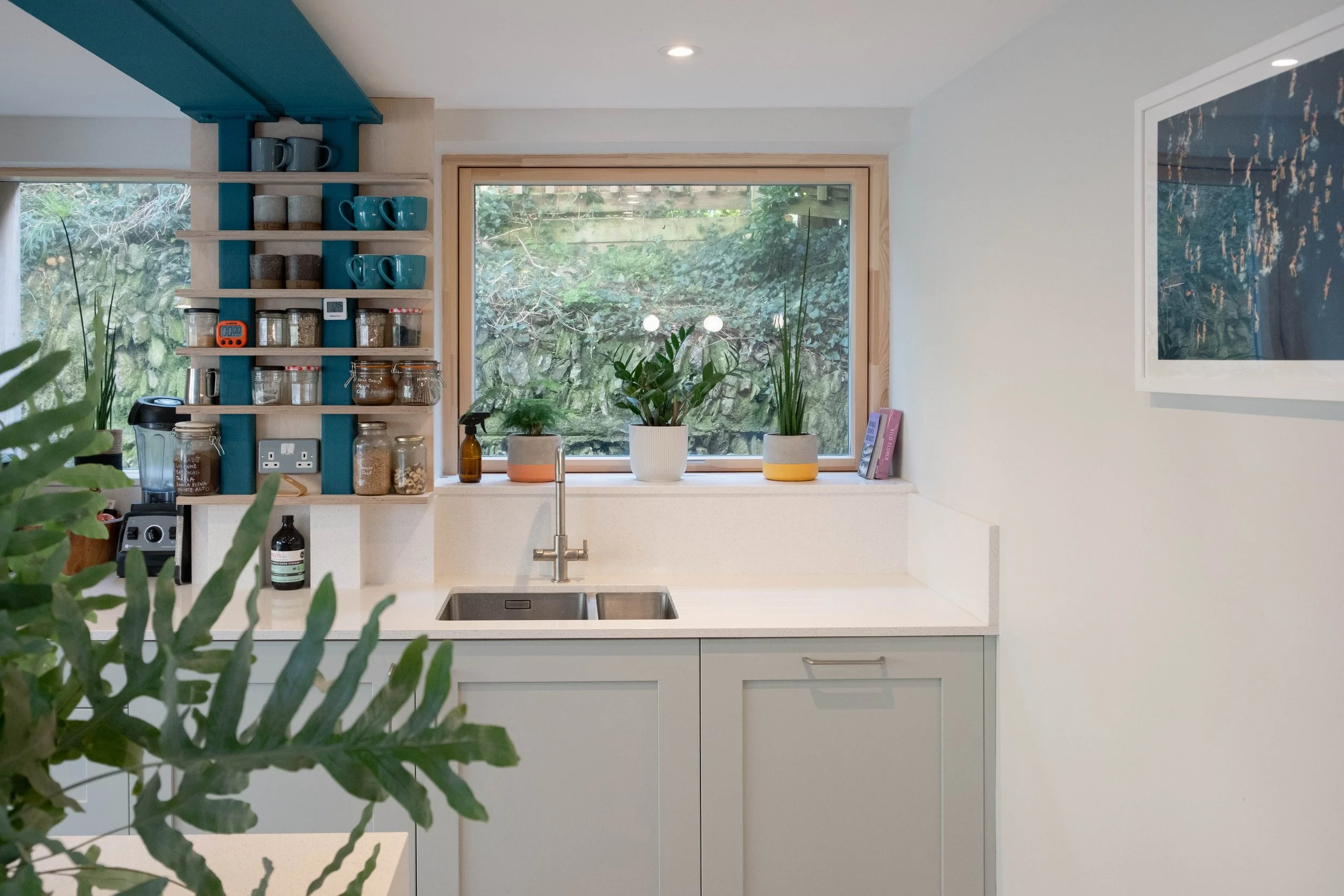 Modern kitchen with a large window overlooking greenery, featuring a white countertop with plants, a cabinet with shelves holding mugs, jars, and bottles, and a framed artwork on the wall.