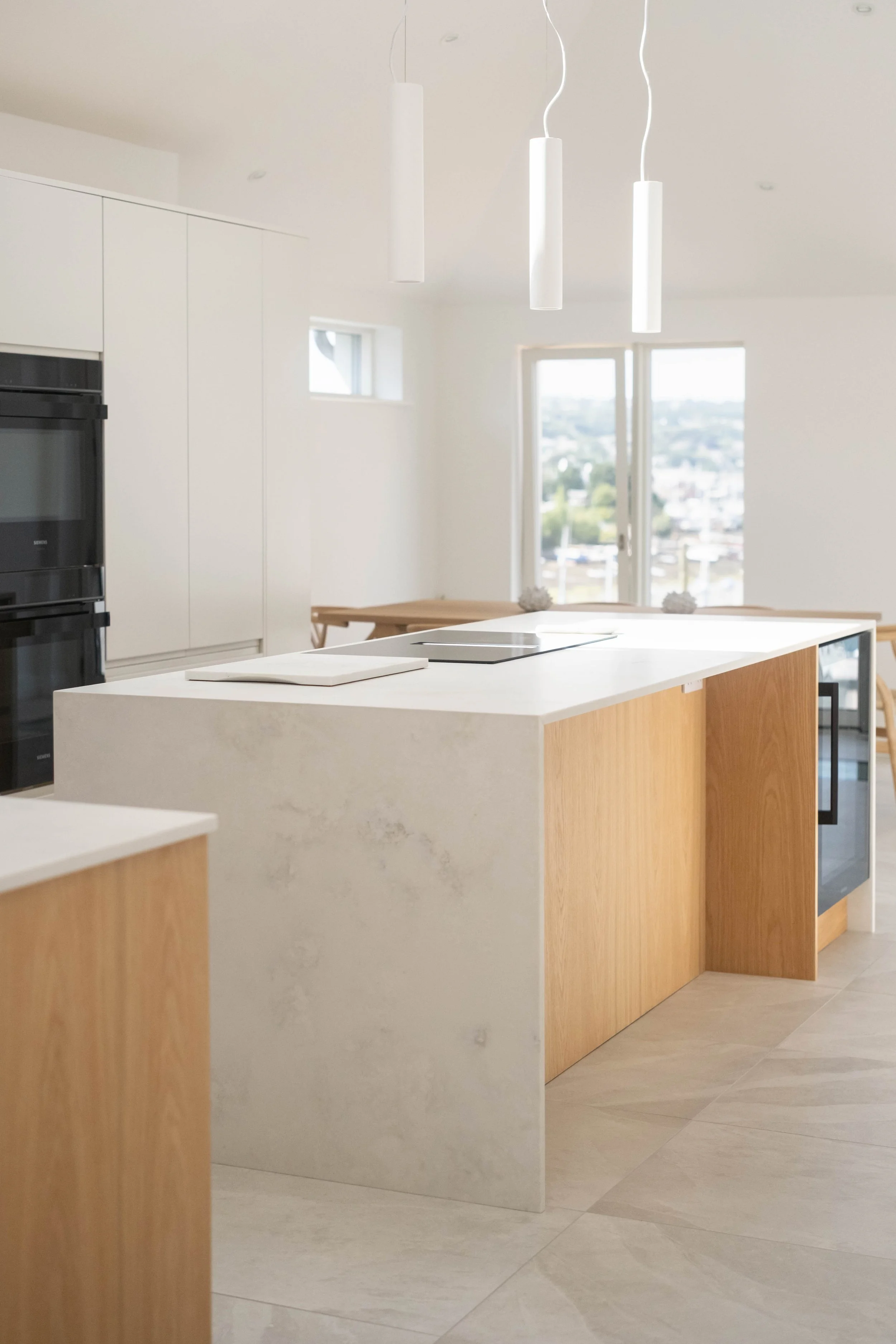 Minimalist kitchen with white countertop, wooden accents, modern pendant lights, and a large window with city views.