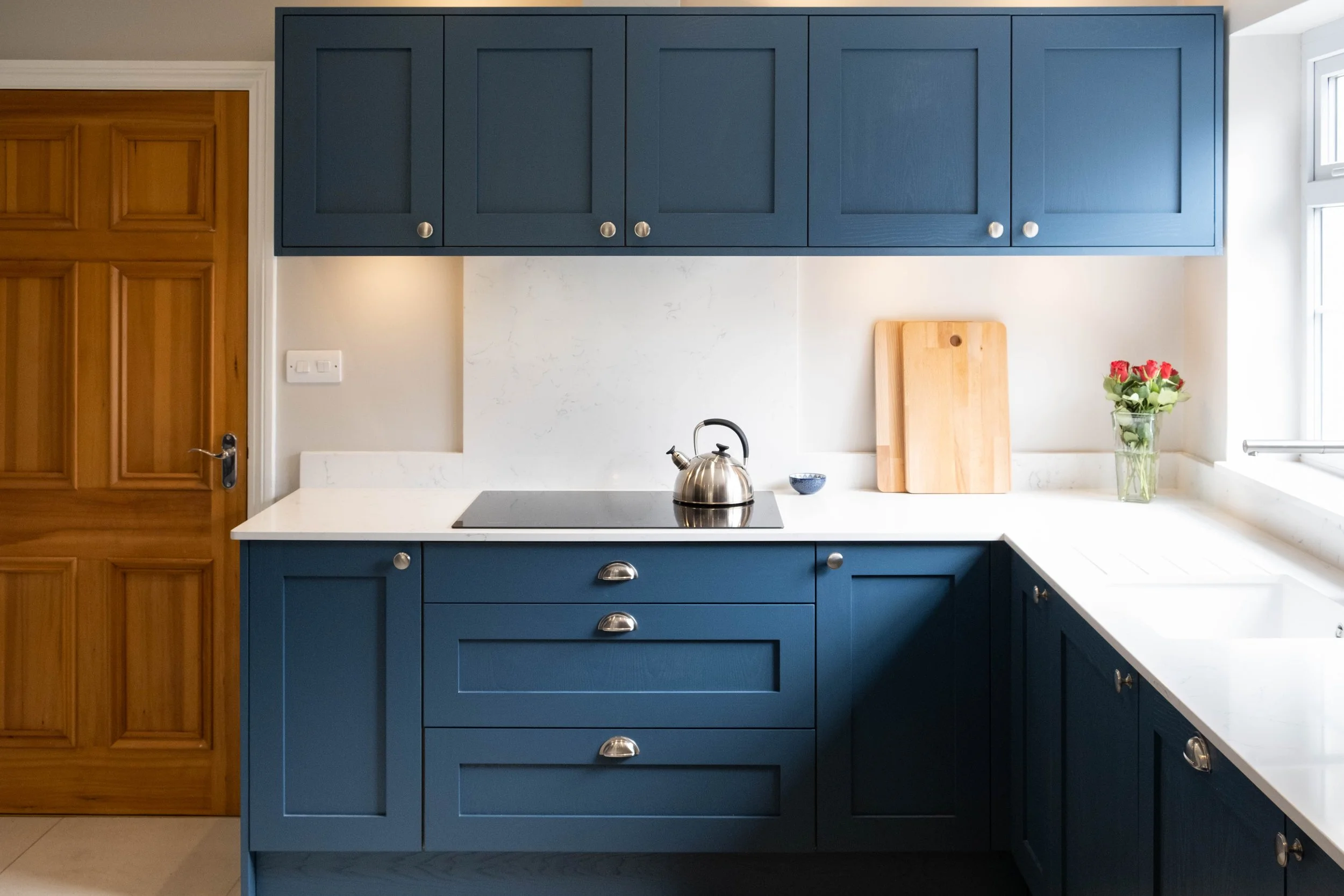 A kitchen with blue cabinets, a white countertop, a wooden door, a small stovetop with a kettle, a vase of roses, and a window to the right.