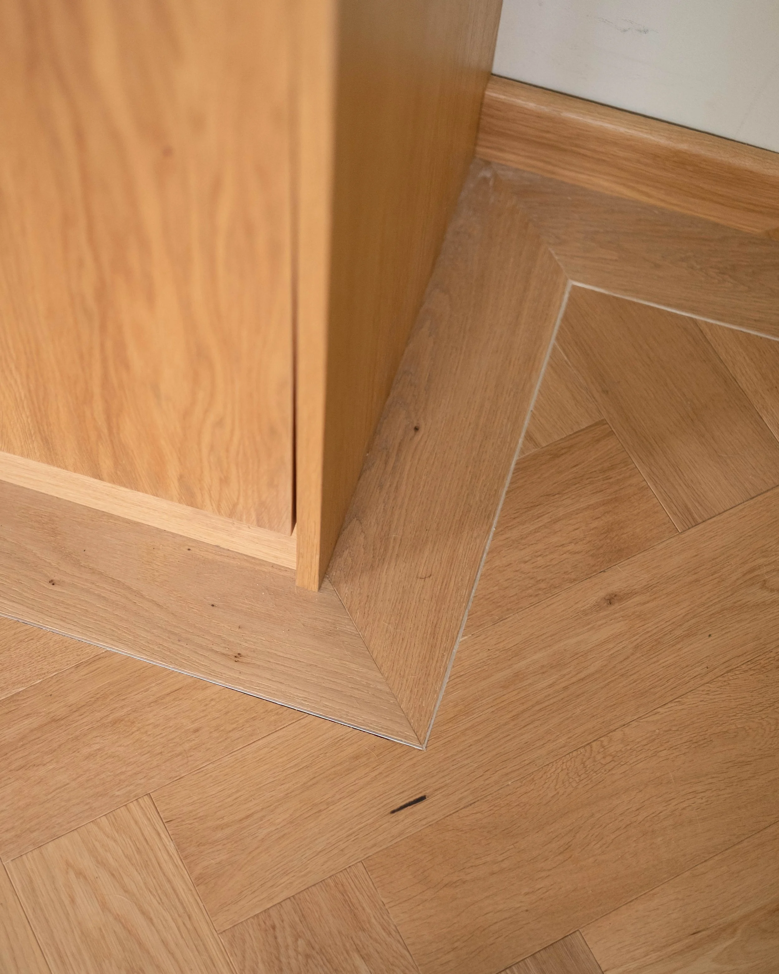 Close-up view of a wooden corner with a floor and wall, showing the grain and texture of the wood, along with some small debris or dirt.