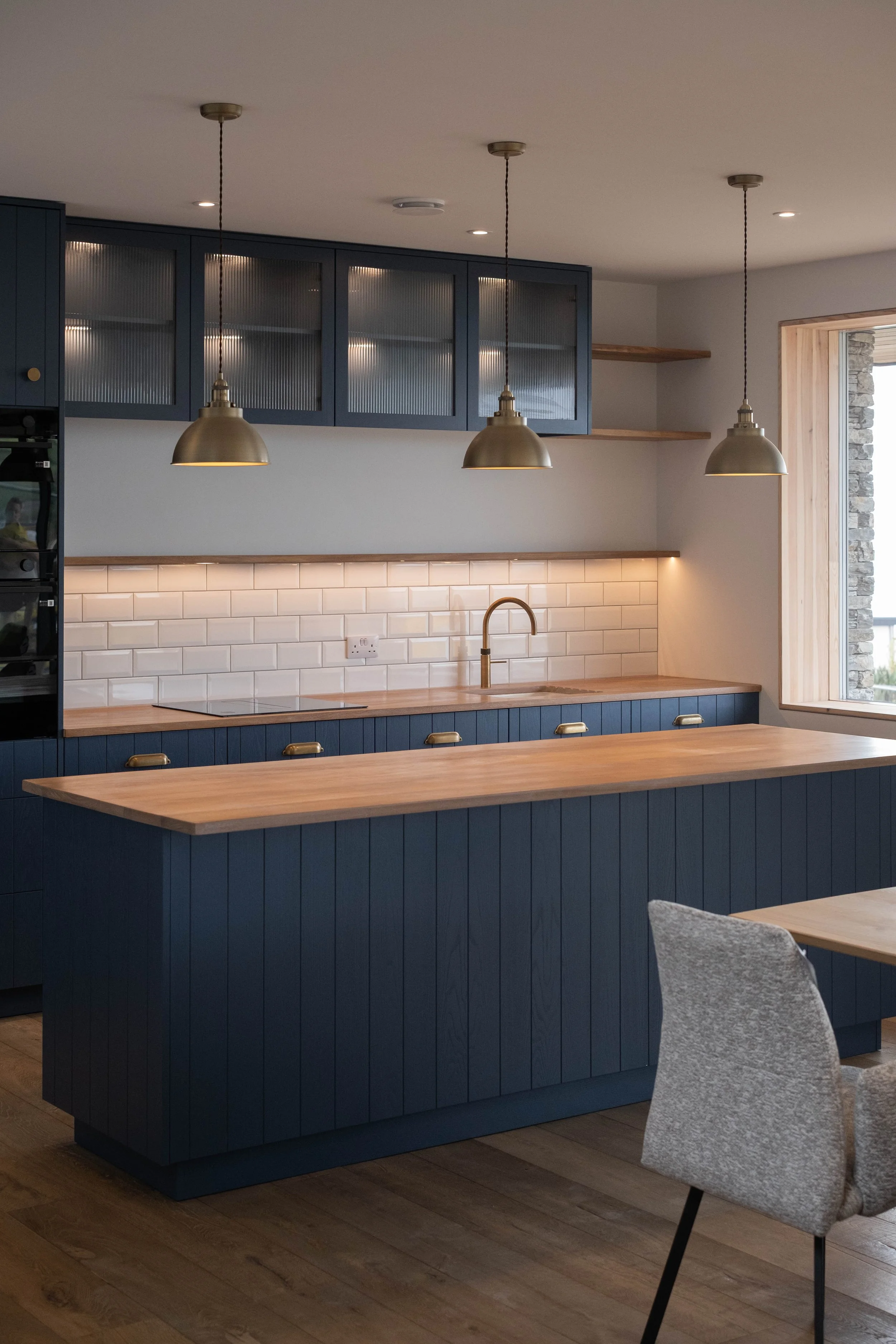 Modern kitchen with navy blue cabinets, wooden countertops, white subway tile backsplash, gold hardware, hanging pendant lights, and a grey upholstered chair.