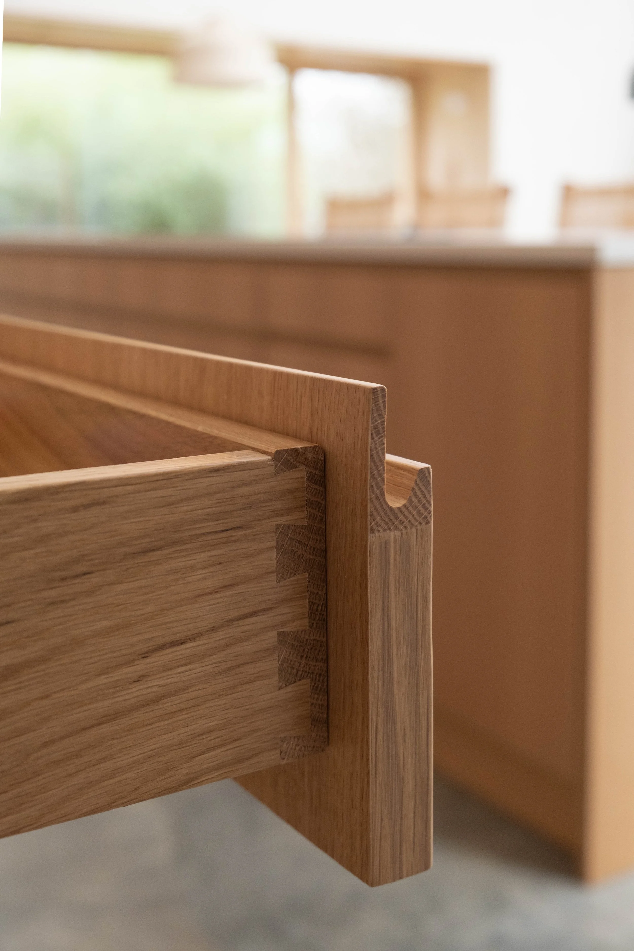 Close-up of wooden furniture with dovetail joints, blurred background of a room with large windows and natural light.