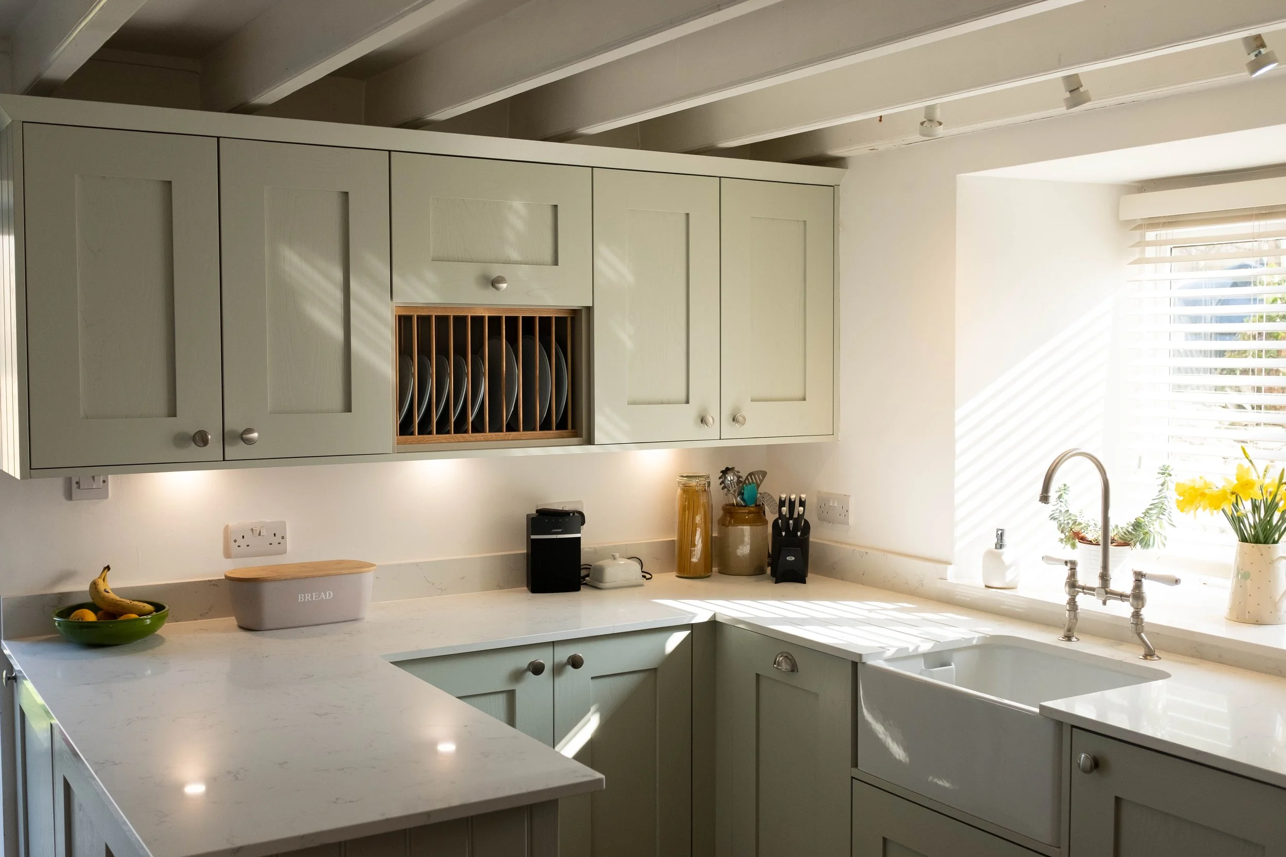 Bright kitchen with green cabinets, white marble countertops, a window with blinds, a white apron front sink, and various kitchen items including a bowl of bananas, bread box, utensils, and decorative plants.