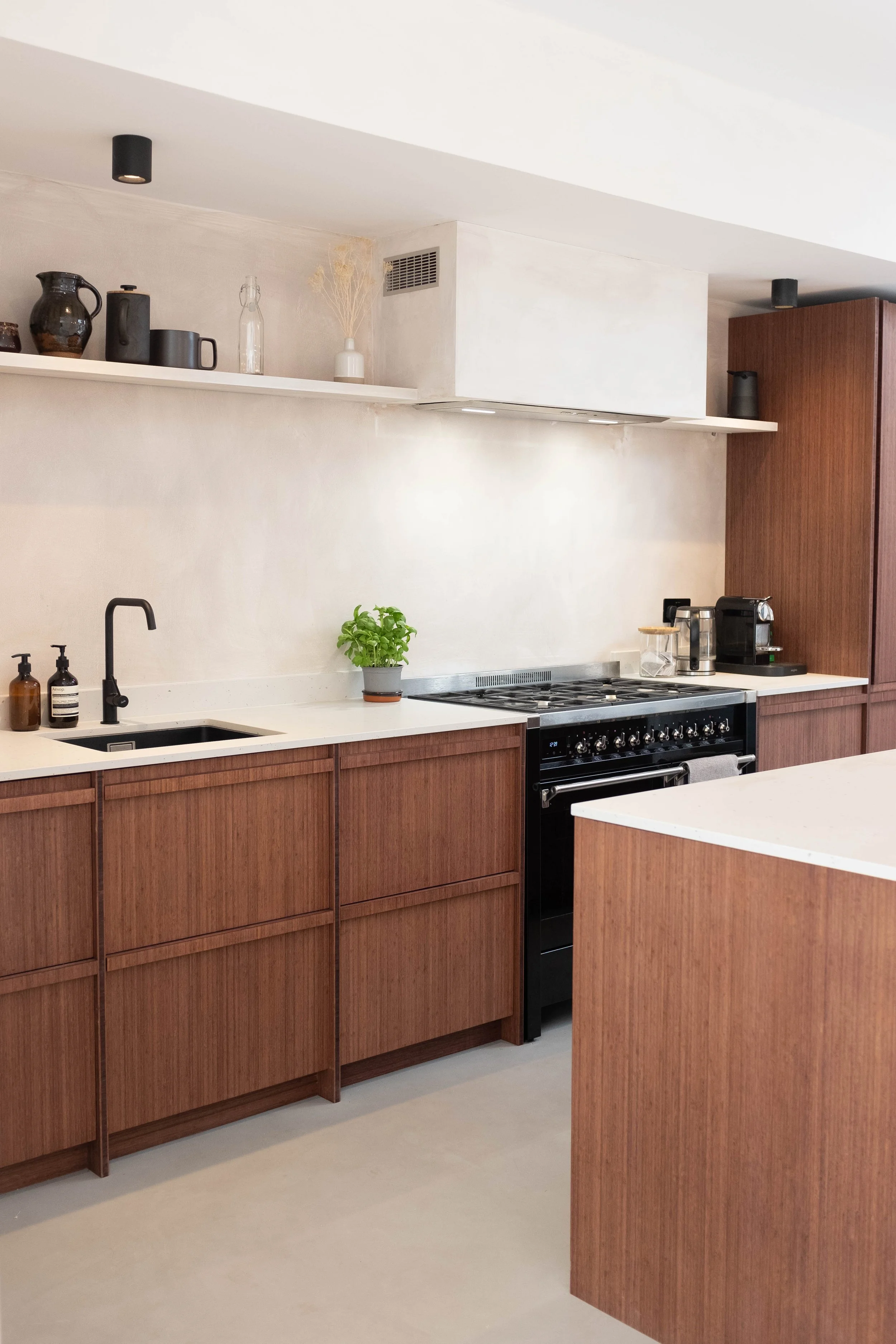 Modern kitchen with wooden cabinets, white countertop, black faucet, potted plant, and appliances including a coffee machine and toaster. Open shelving displays black dishware and decorative items.