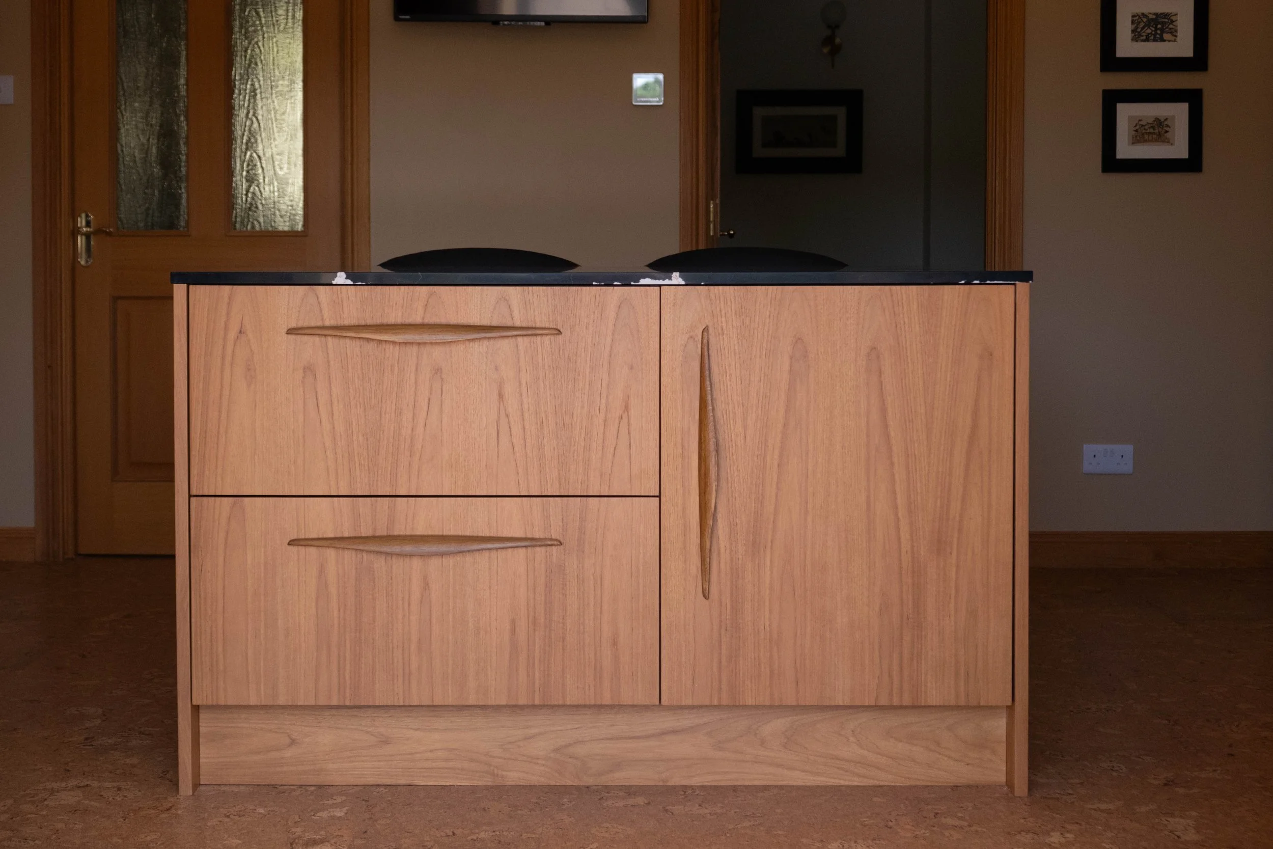 A wooden cabinet with three drawers and two doors, topped with a black countertop, situated in a room with beige walls and wooden trim. There is a double wooden door with glass panels to the left and framed pictures on the walls.