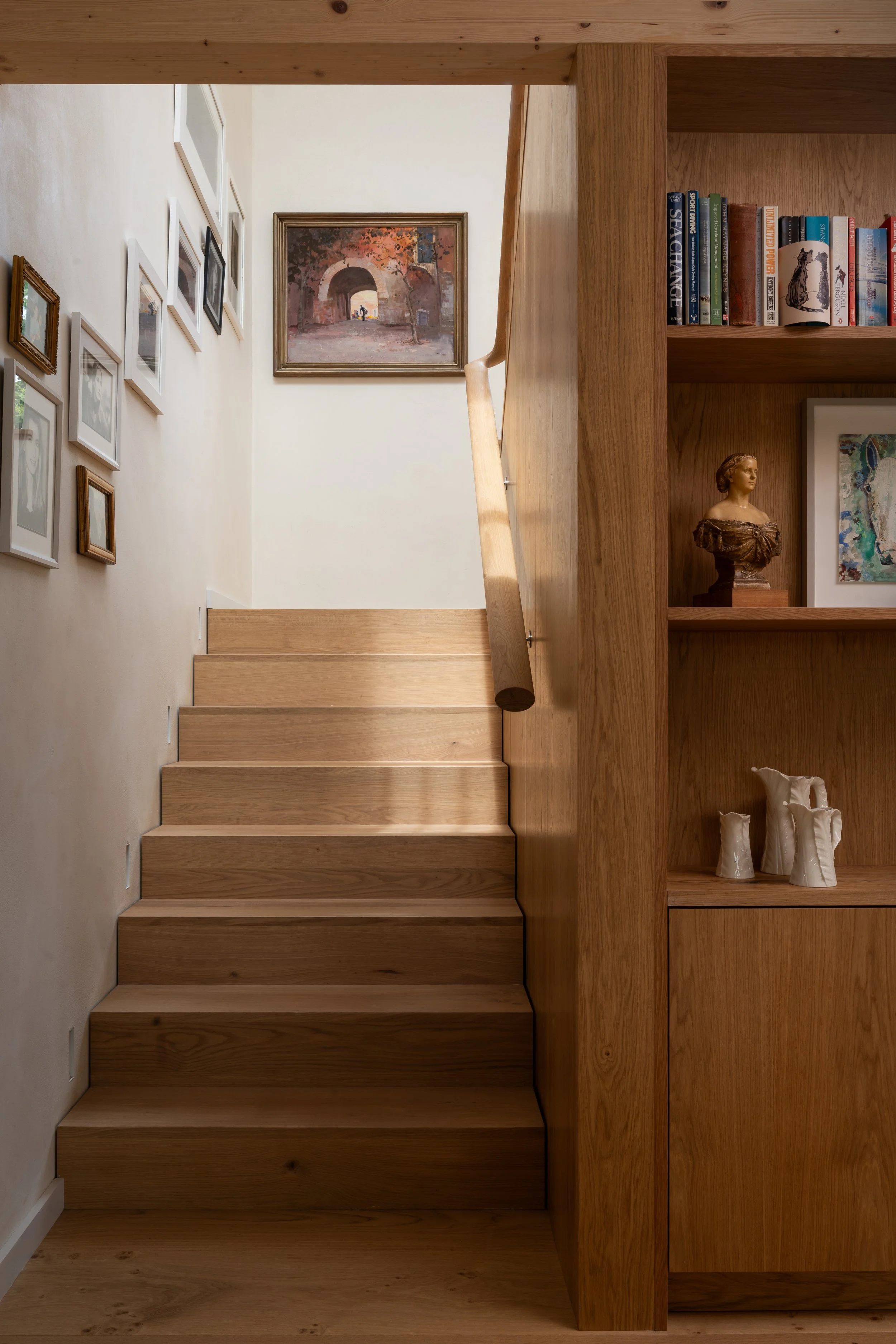 Interior view of a wooden staircase next to a built-in bookshelf with books, a bust sculpture, and vases, with framed artwork on the wall.