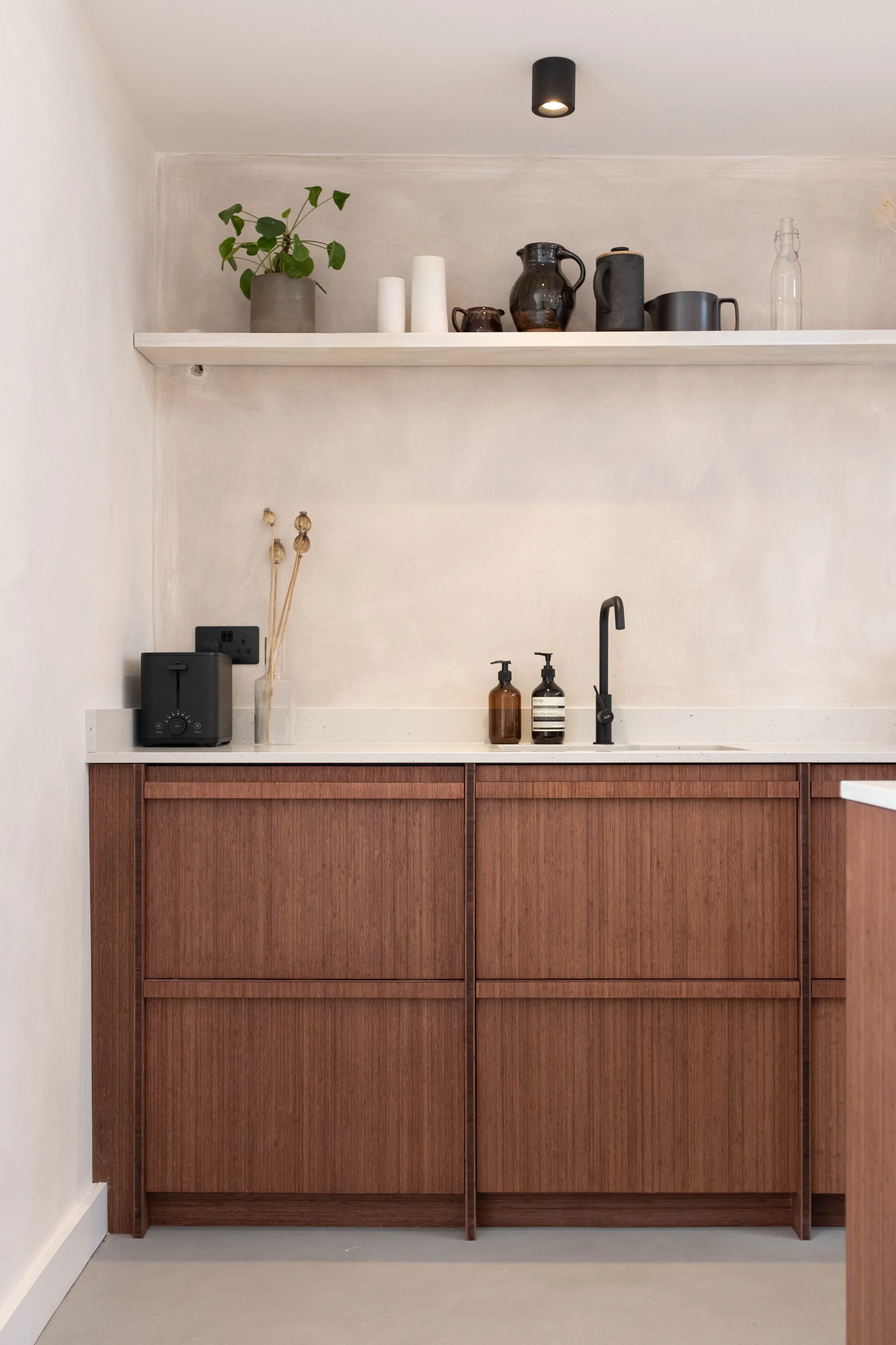 Kitchen counter with two brown cabinets, a white countertop, two bottles of soap, a black faucet, and a wall socket. Above the counter, a white shelf holds a potted plant, white vases, dark pottery, a glass bottle, and a black light fixture.