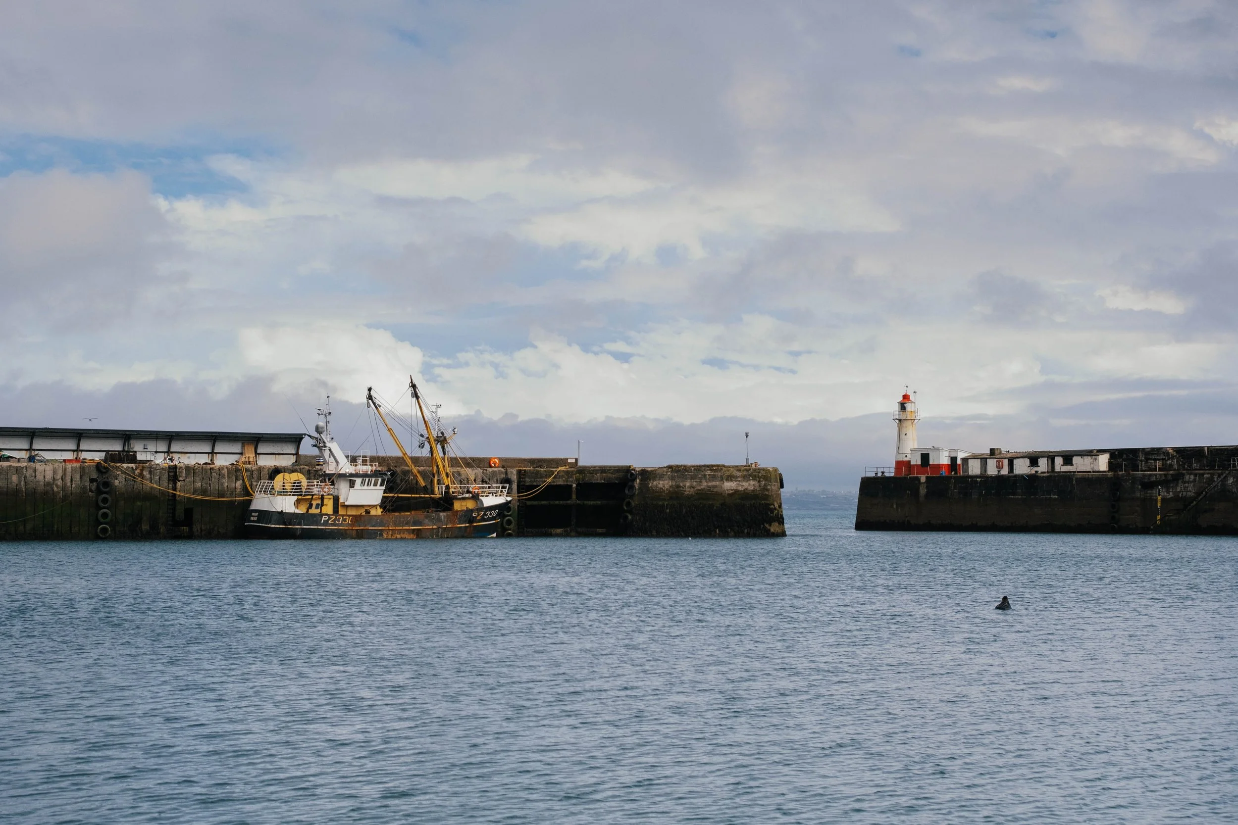 A harbor scene with a small boat docked alongside a concrete pier, a lighthouse in the background, and a cloudy sky overhead.