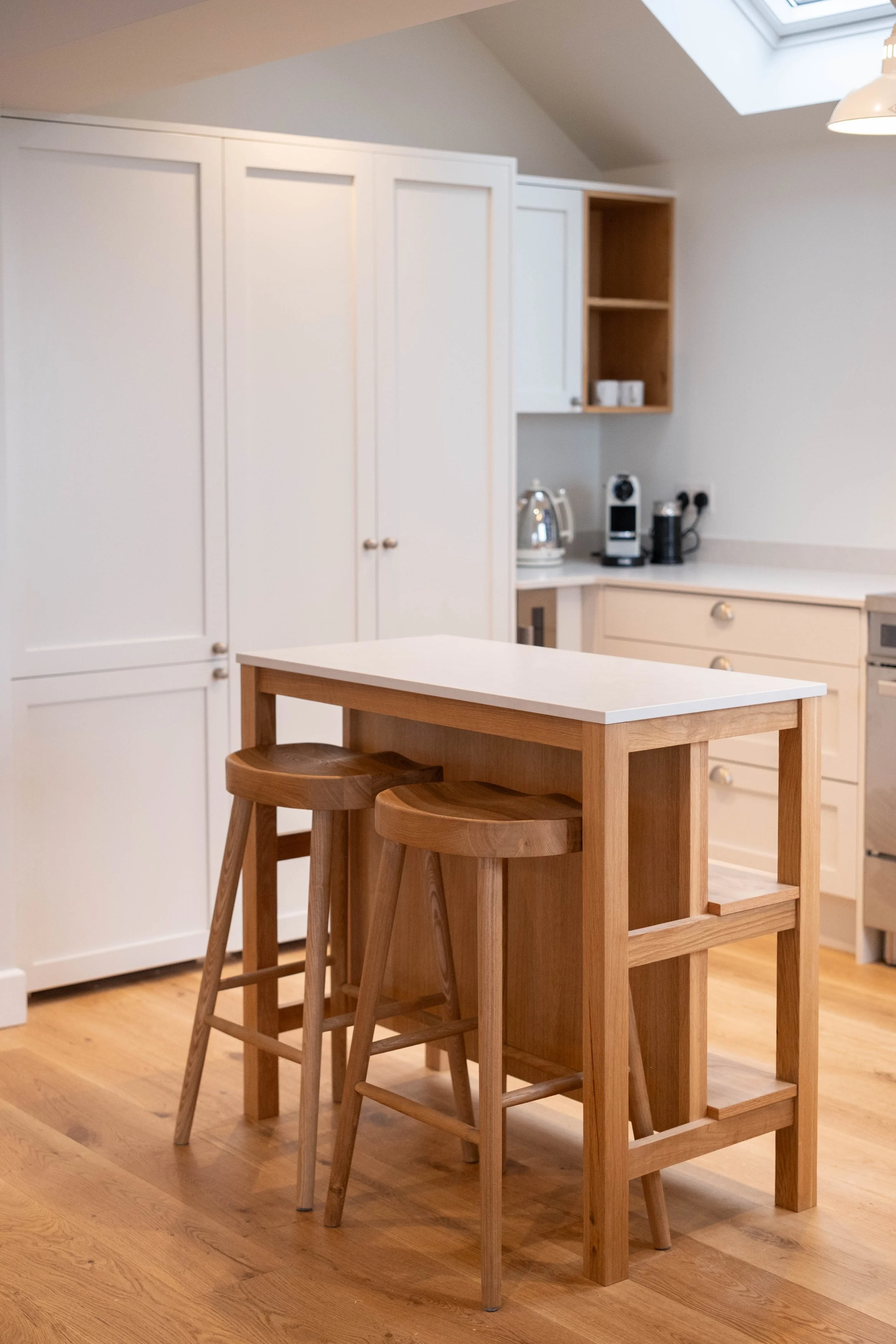 Wooden kitchen table with three matching wooden bar stools in a modern kitchen with white cabinets, a kettle, and coffee maker on the countertop, and natural light coming from a skylight.