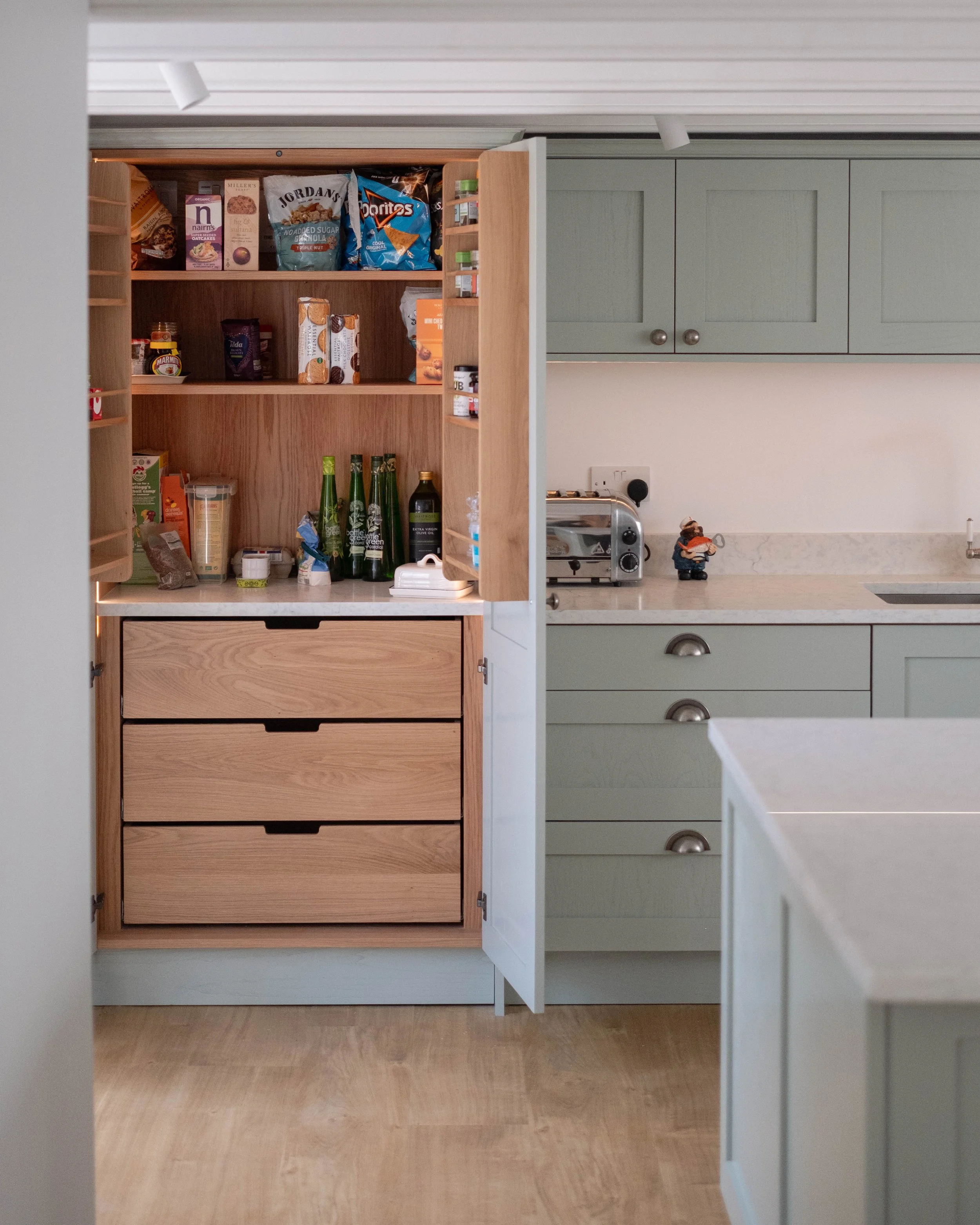A kitchen pantry with an open wooden cabinet filled with food items on shelves, and a separate section with green cabinets and a countertop with small appliances and decorations.