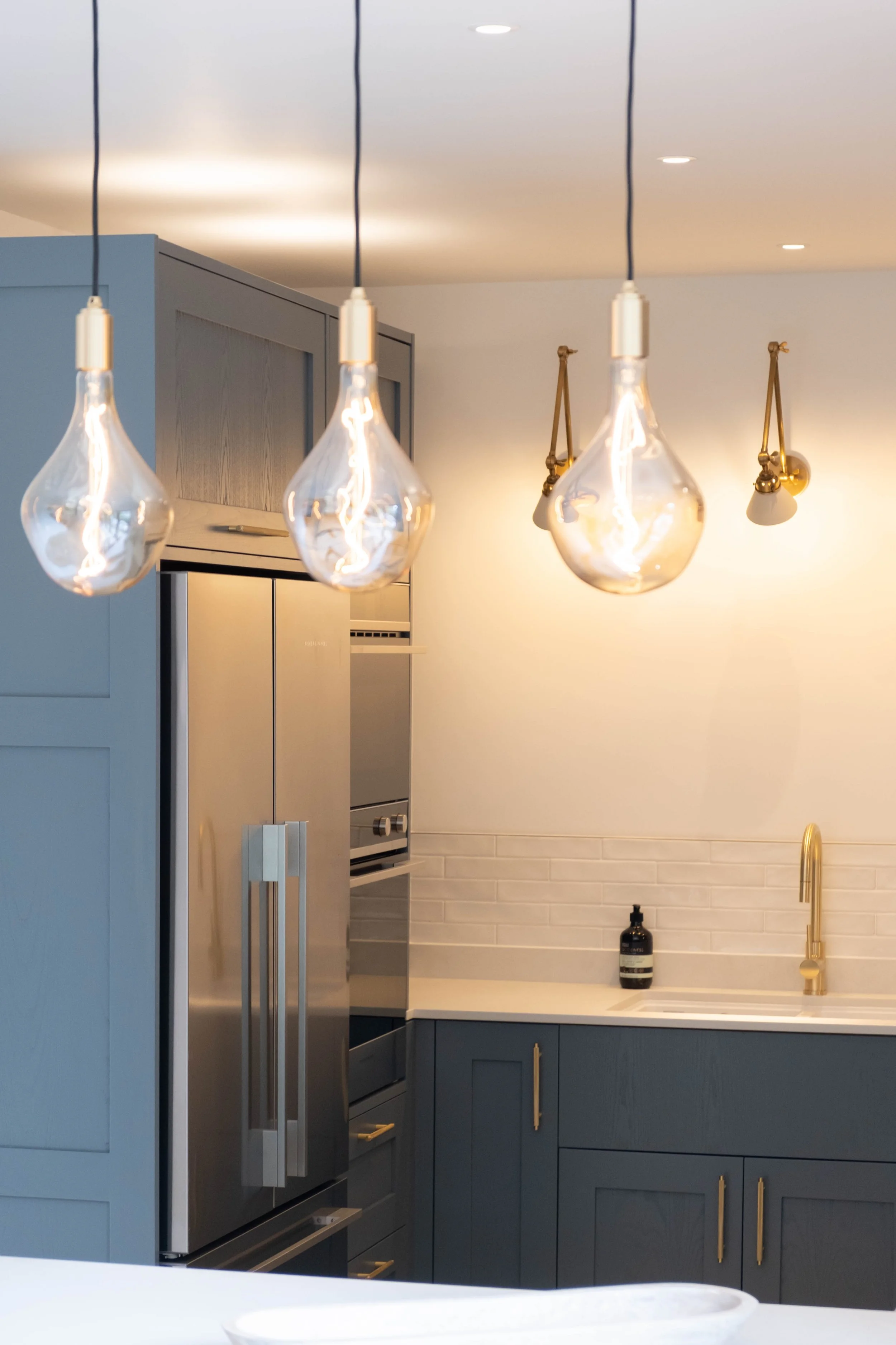 Modern kitchen with blue cabinets, stainless steel refrigerator, white countertop, gold handles, white subway tile backsplash, black soap dispenser, sink, gold faucet, hanging pendant lights with exposed bulbs, and ceiling spotlights.