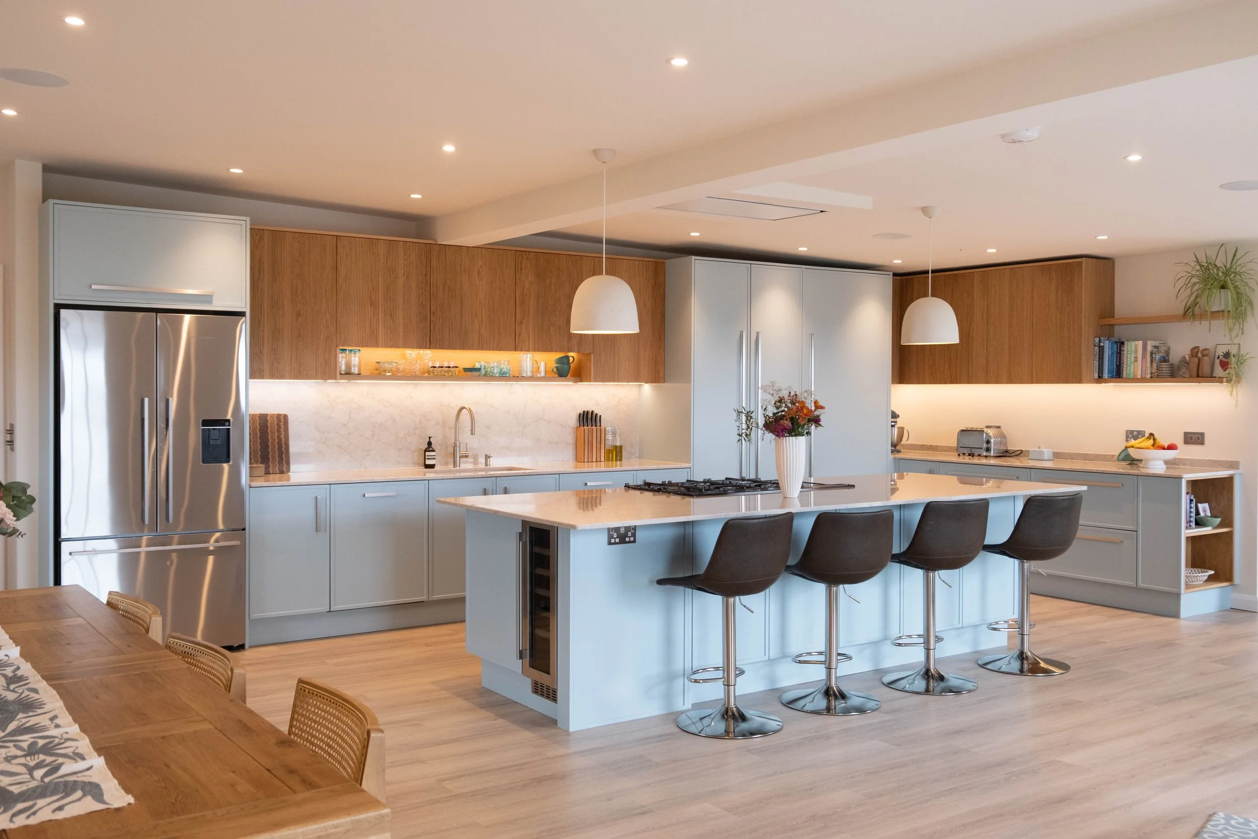 Modern kitchen with white and wood cabinets, stainless steel appliances, a kitchen island with four bar stools, pendant lighting, and open shelving with books and plants.