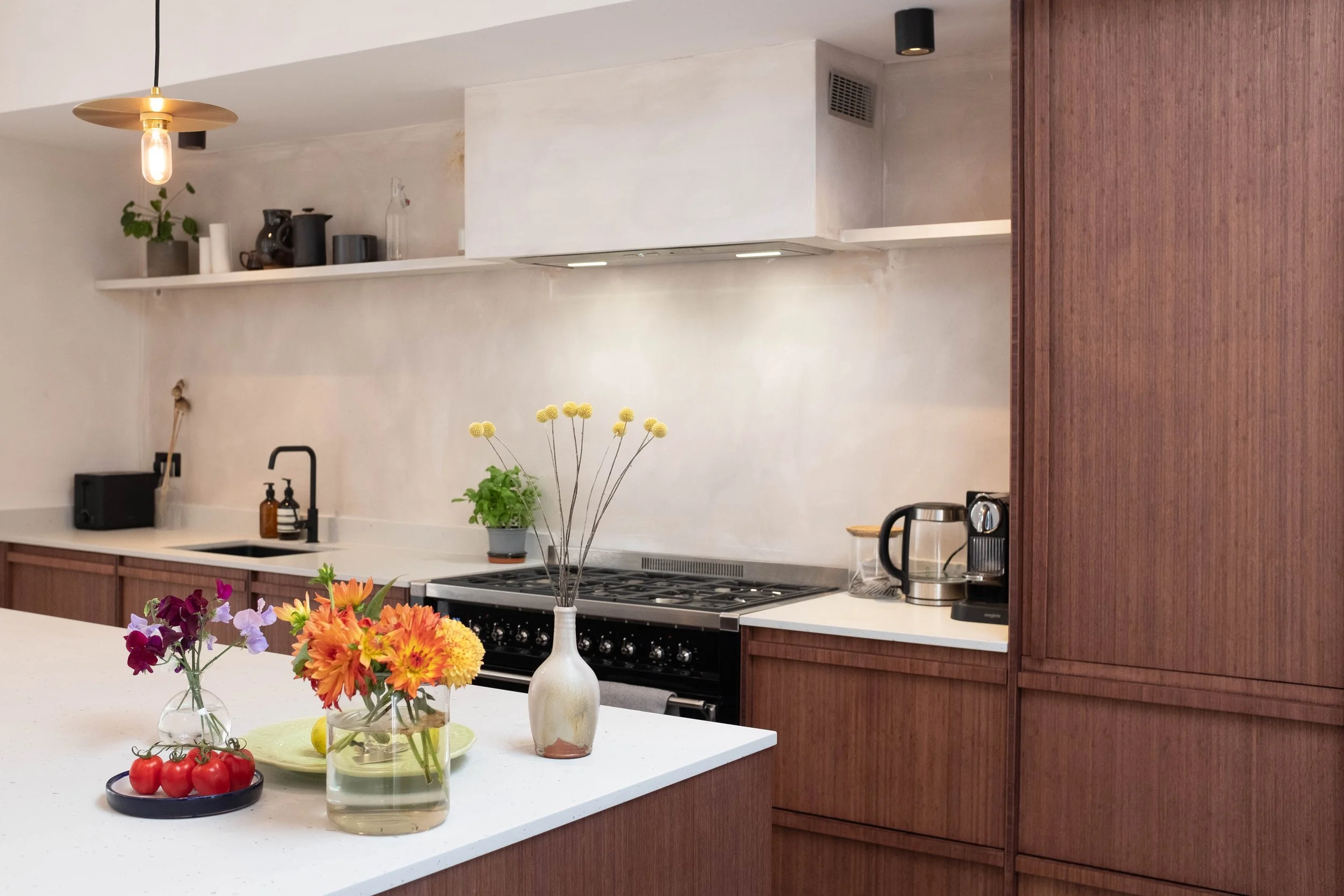 Kitchen with a white countertop, wooden cabinets, and a black stove. Decorated with flowers in a vase, tomatoes, a potted plant, and kitchen appliances.
