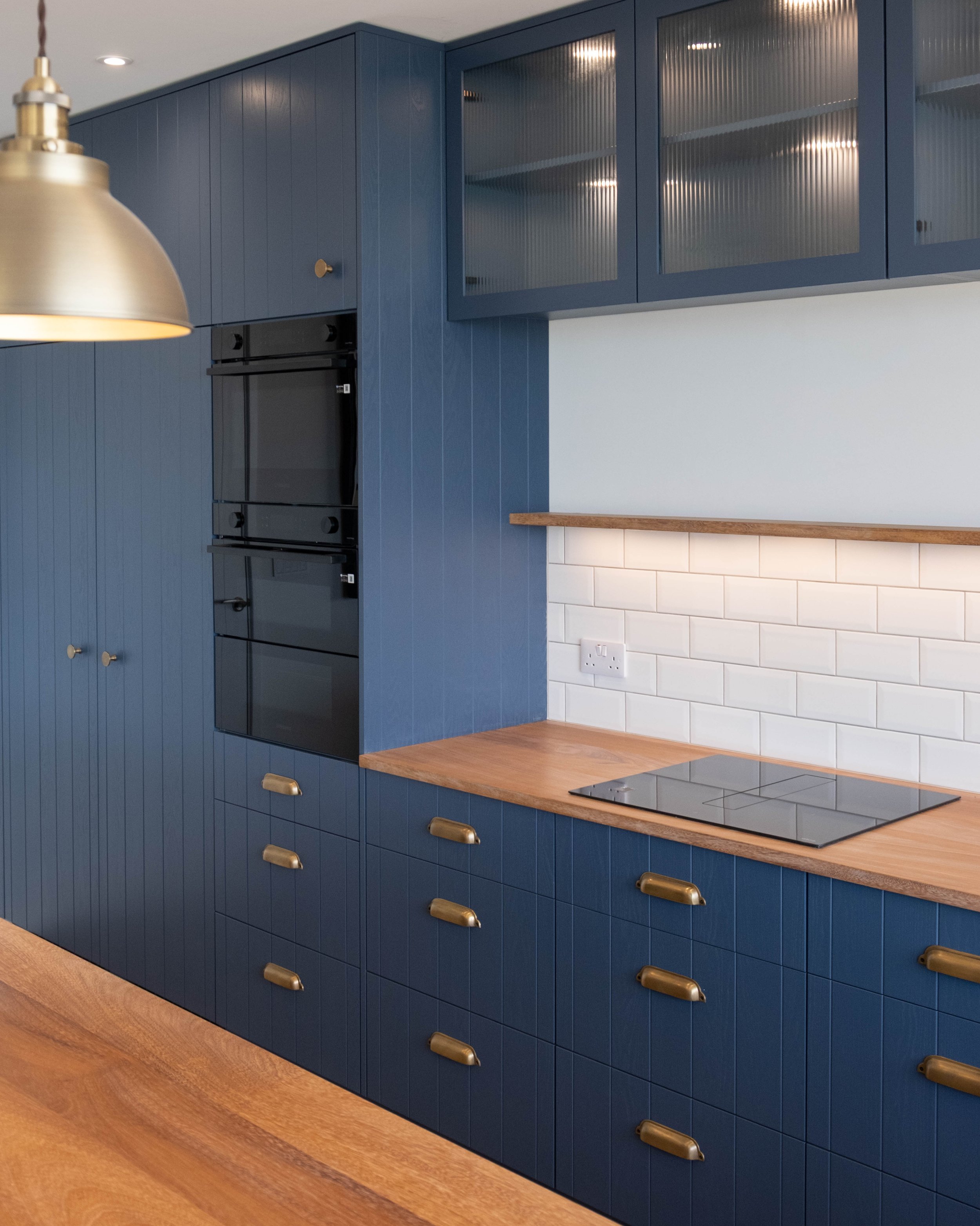 Modern kitchen with blue cabinetry, wooden countertop, white tile backsplash, black oven, and gold handles and fixtures.