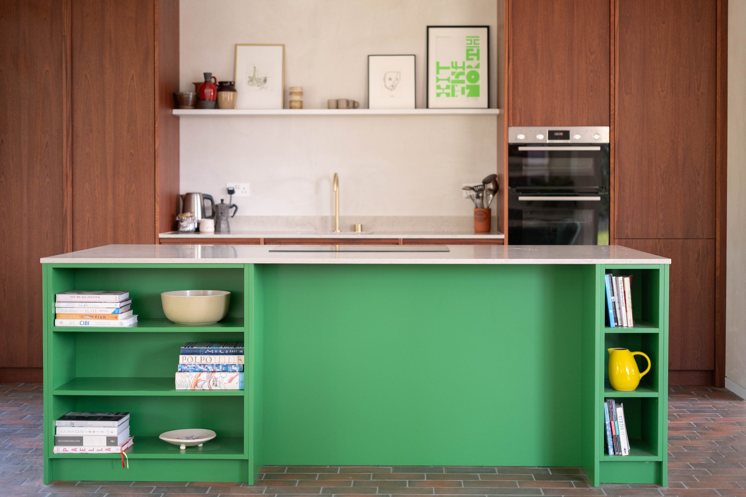 A modern kitchen with a white marble countertop, wooden cabinets, a built-in oven, and a green bookshelf island holding books and decorative items.
