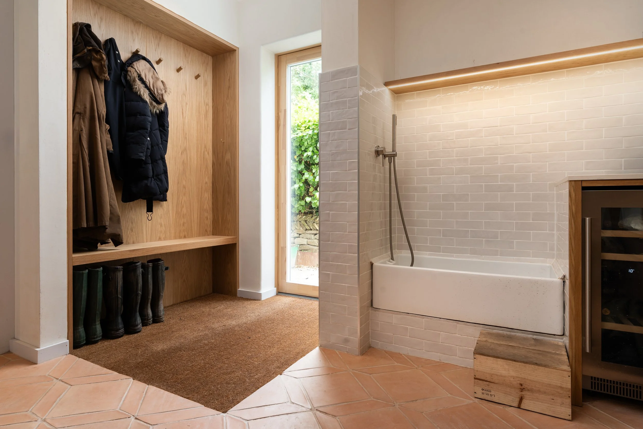 Entryway with coat hooks and shoes on a wooden bench, leading to a bathroom with a bathtub and shower, tiled walls, and a small refrigerator.