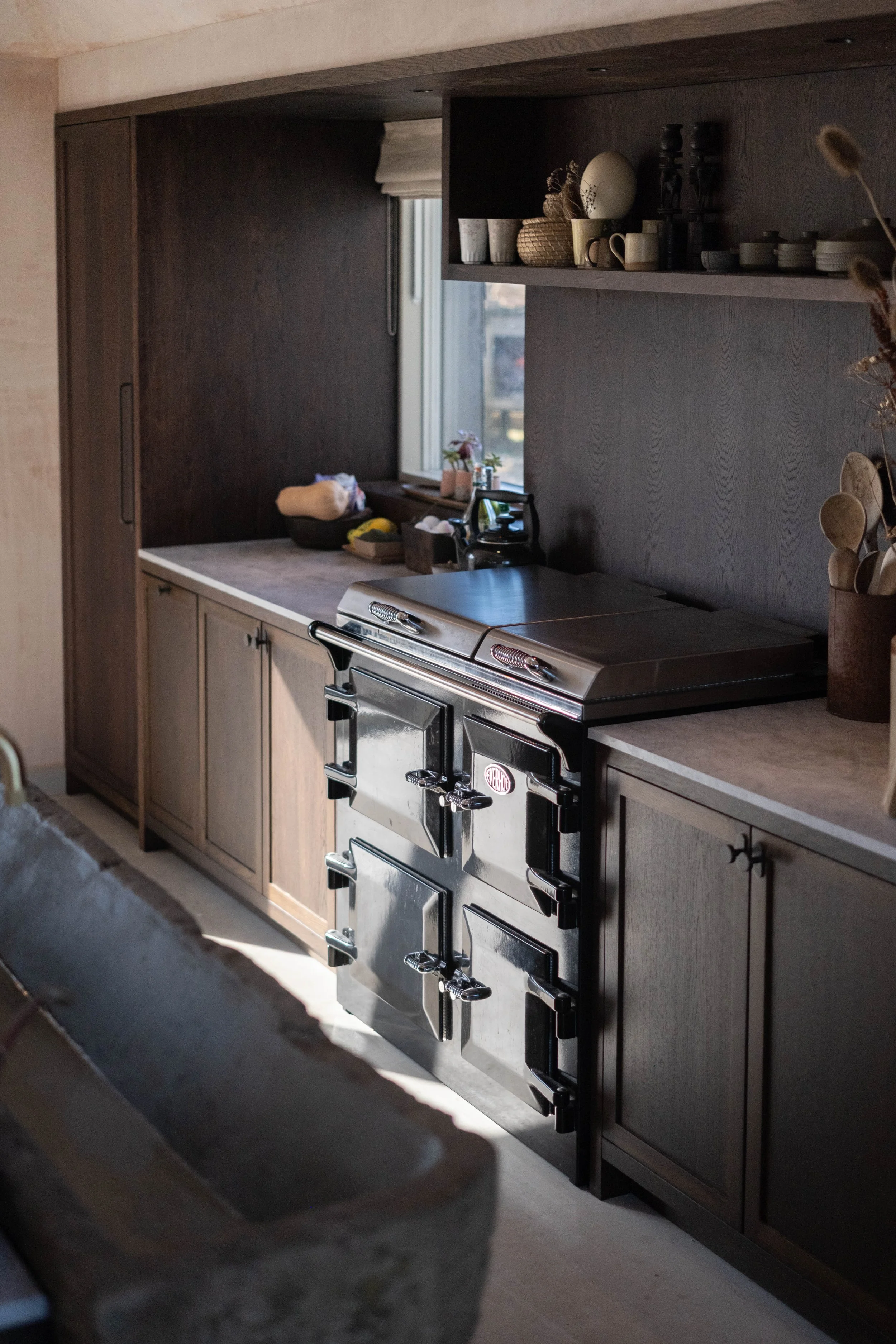 A kitchen with dark wood upper cabinets and light wood lower cabinets, a stove, a window with a view outside, and various kitchen items on the countertop and shelves.