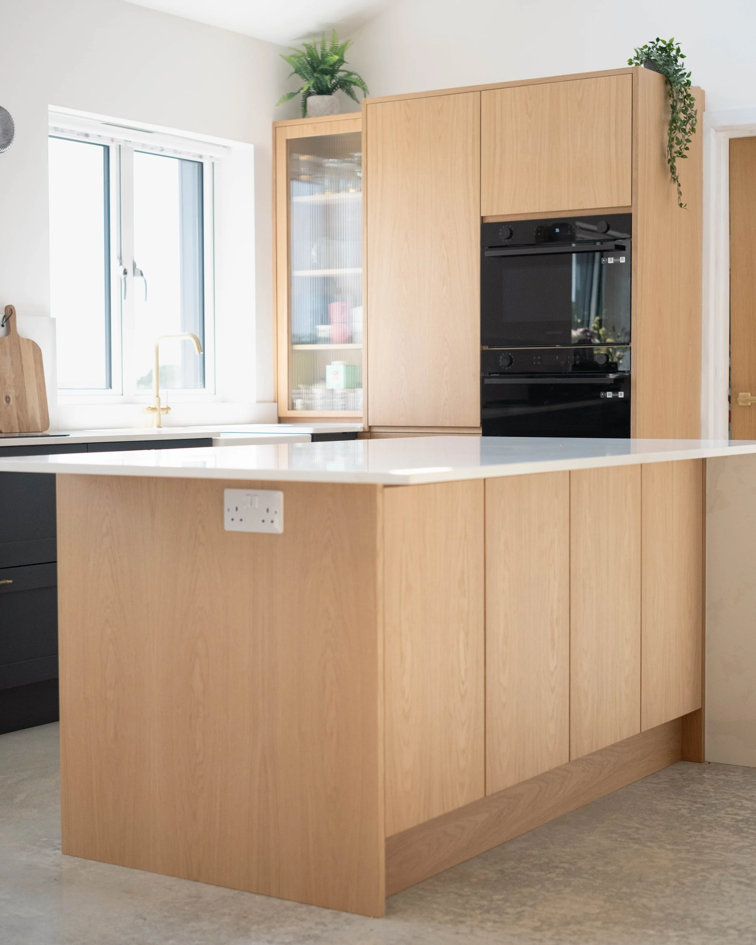 A modern kitchen with light wood cabinetry, a white countertop, a window with a blind, and green plants on top of the cabinets.