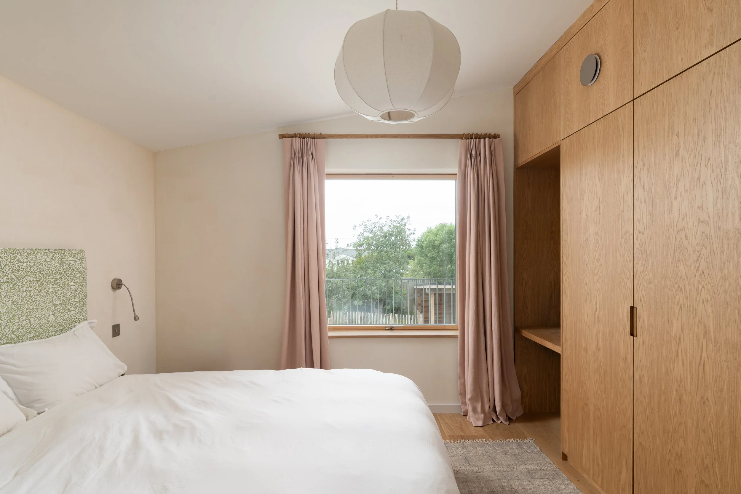 A bedroom with a bed, white bedding, a green patterned headboard, a wall-mounted reading lamp, a window with pink curtains, and a wooden cabinet or wardrobe.