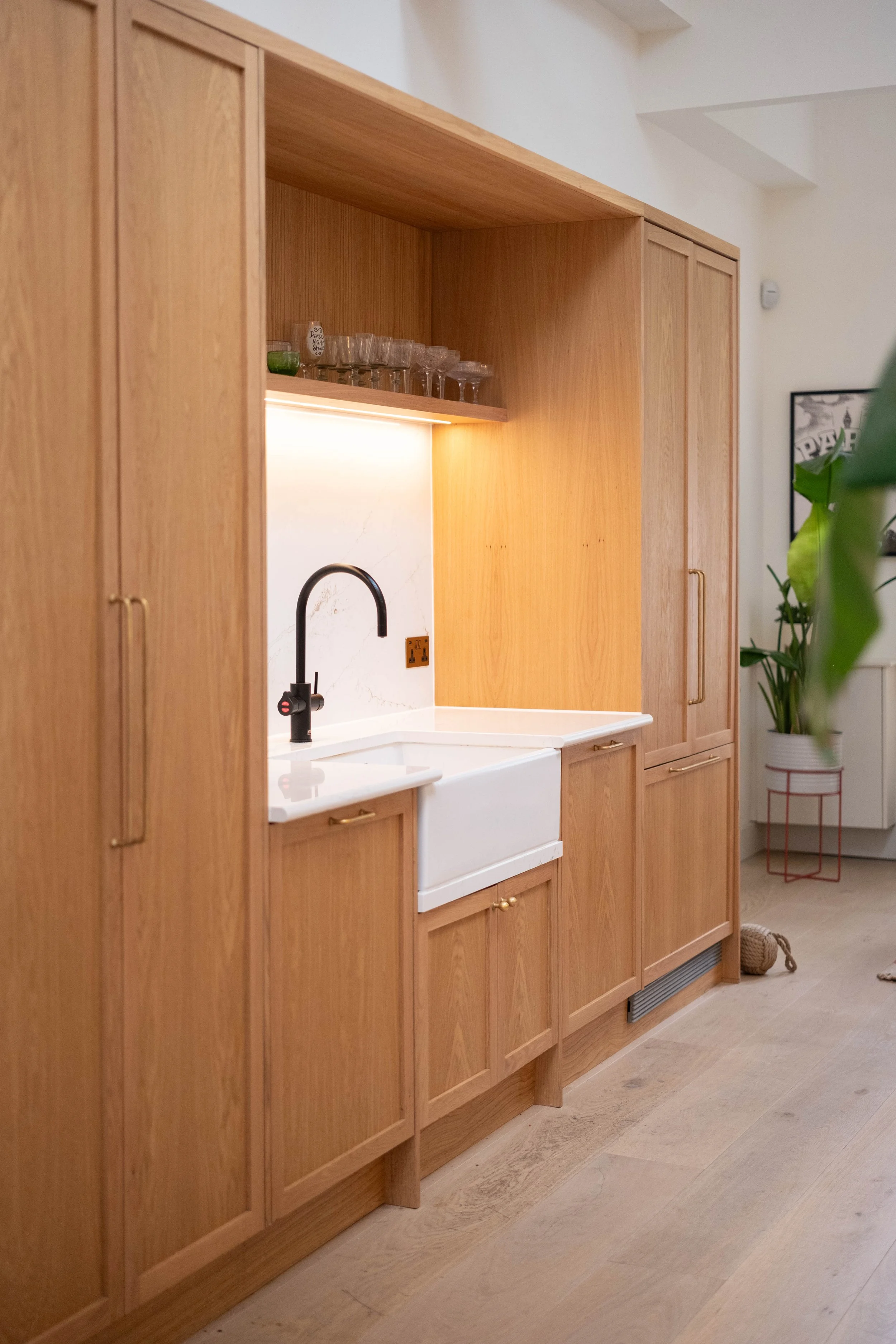 A kitchen with wooden cabinetry, a white farmhouse sink, and a black faucet. There are glasses on a shelf above the sink, and a plant in a pot nearby. The floor is light-colored wood.