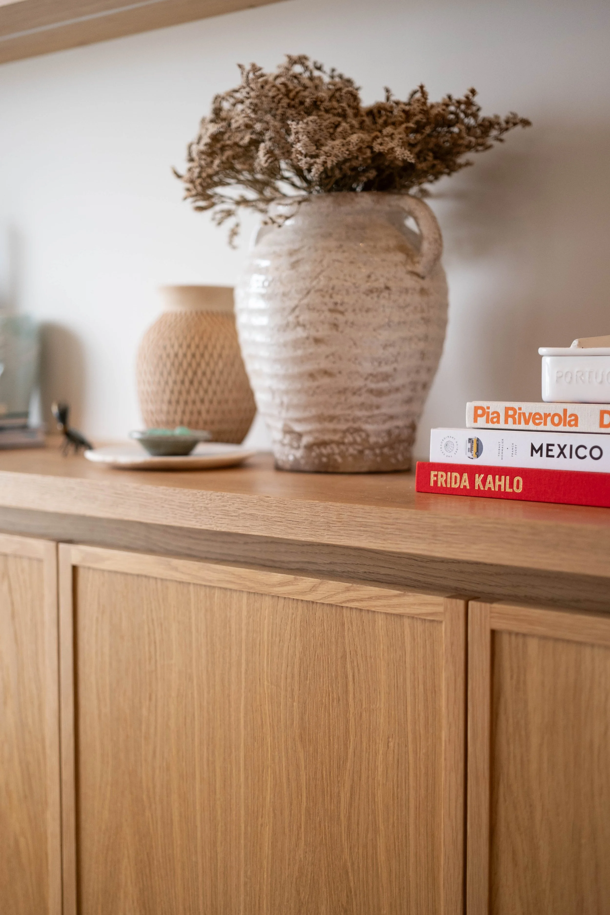 A light-colored wooden sideboard or cabinet with a large ceramic vase containing dried flowers on top, and a stack of books beside it.