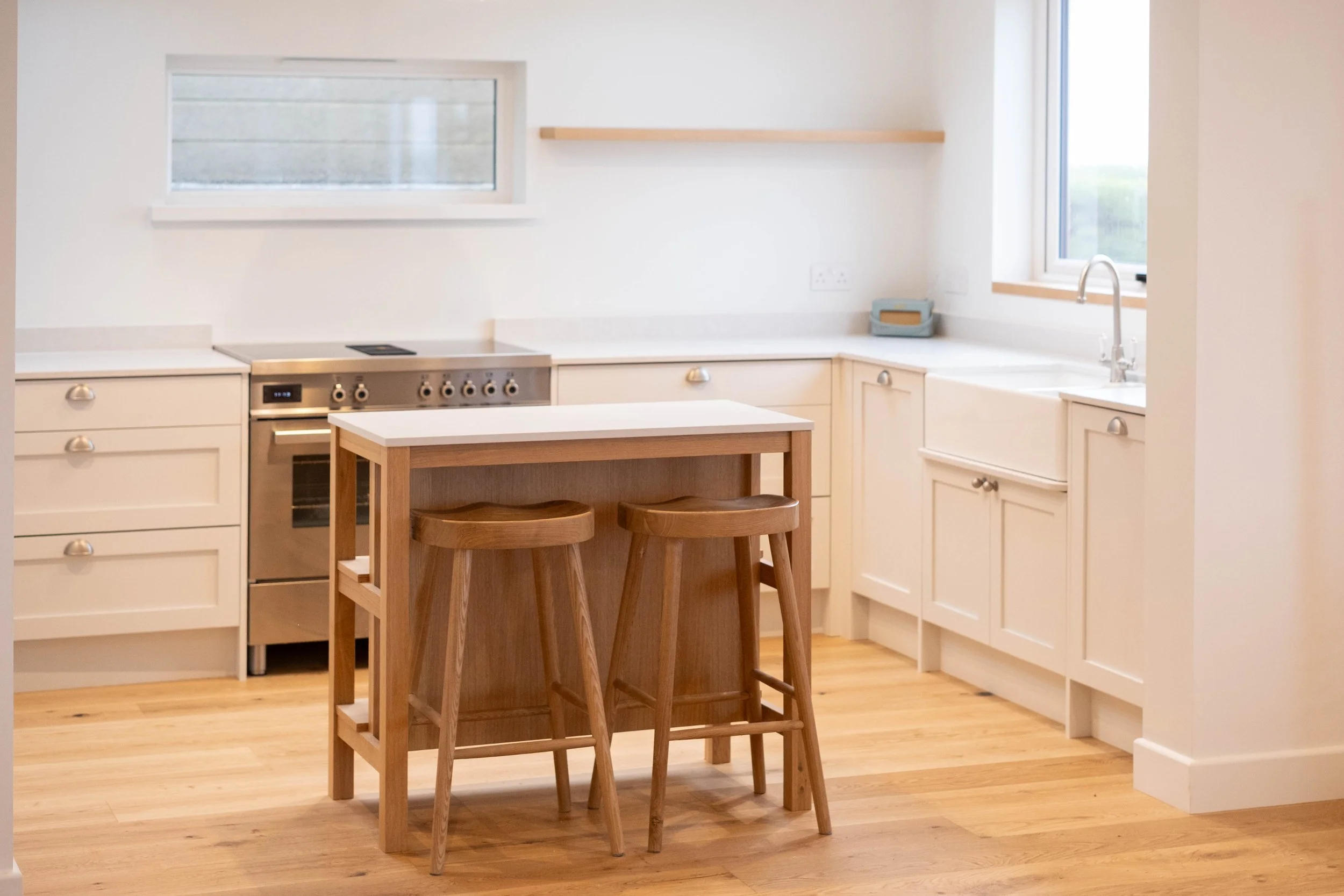 A modern kitchen with white cabinets, a wooden island with two bar stools, a stainless steel oven with control knobs, two windows letting in natural light, and a wooden floor.