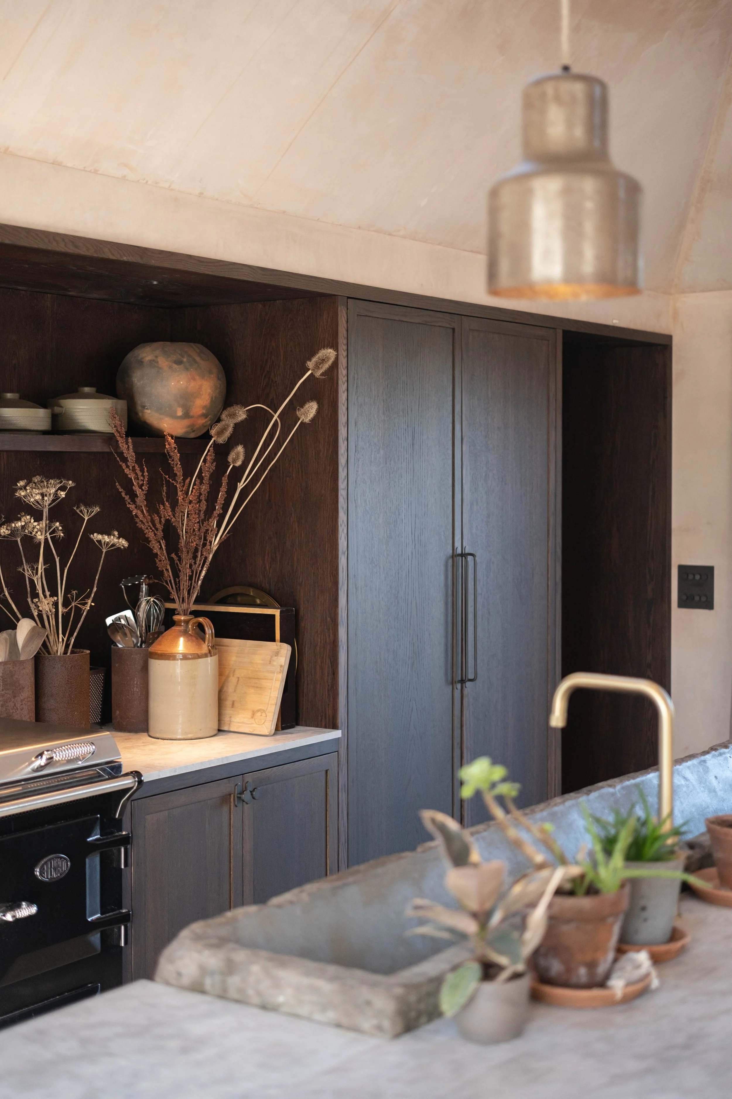 Kitchen with dark wooden cabinets, a marble countertop with potted plants, a ceramic vase, dried flowers, and a glass jar, with a visible oven and a modern light fixture hanging from the ceiling.