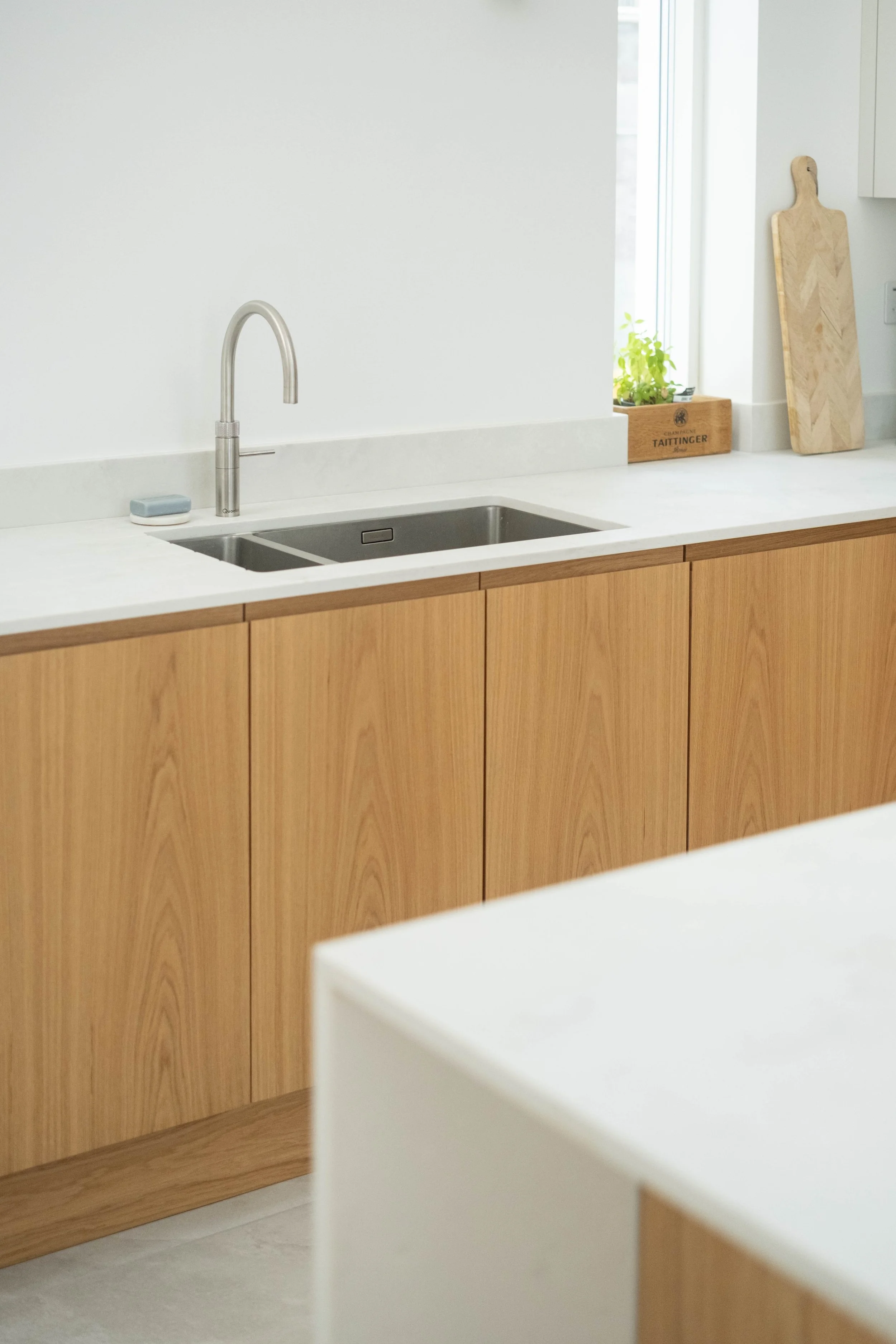 A domestic kitchen with light wood cabinets, a white countertop, a stainless steel sink, a scrubber, a small plant near the window, and a wooden cutting board.