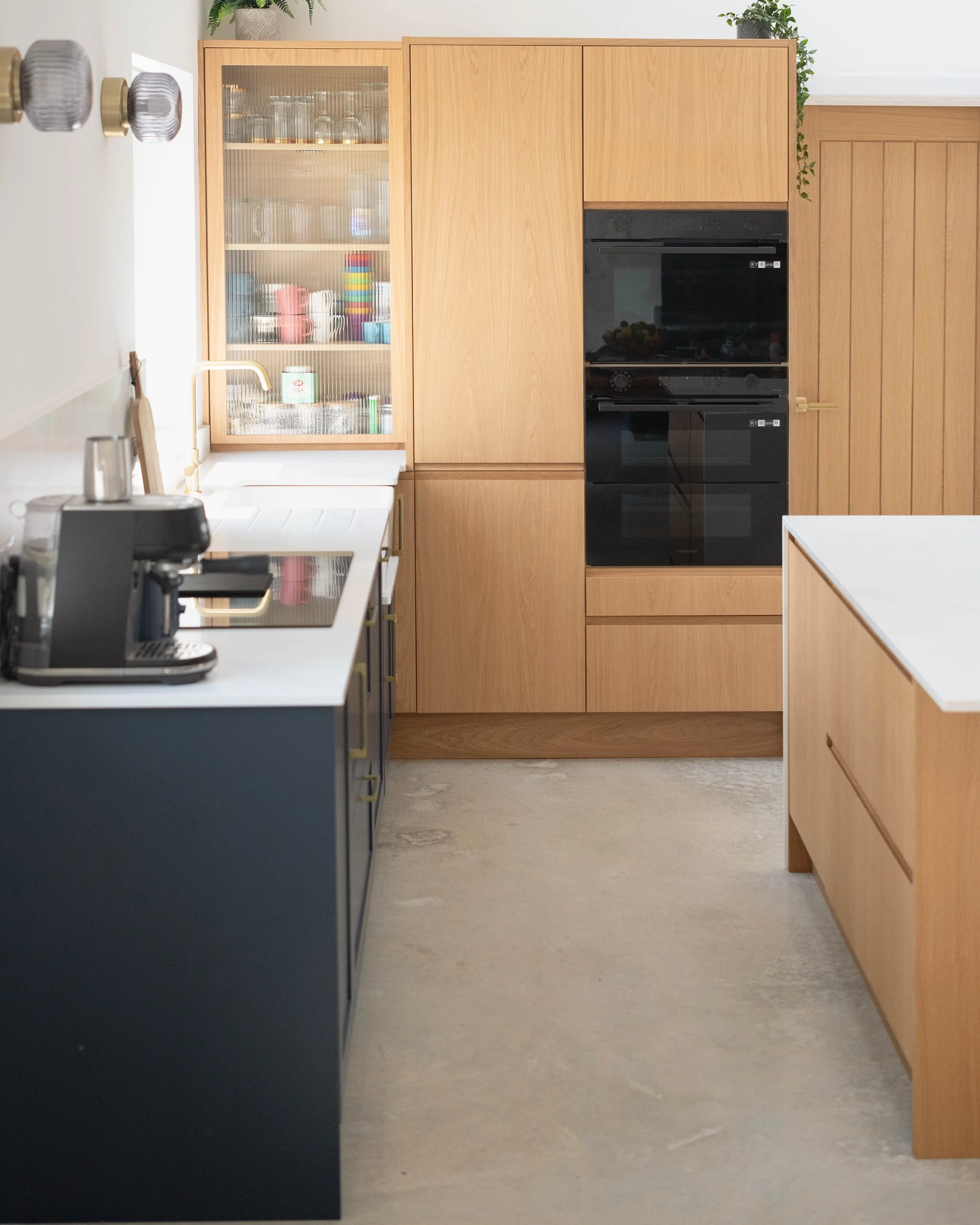 A modern kitchen with light wood cabinets, a white countertop, and a black oven built into the cabinets. There is a glass-front cabinet filled with glassware and colorful mugs, and some plants on top of the cabinets. On the left side, part of a kitch
