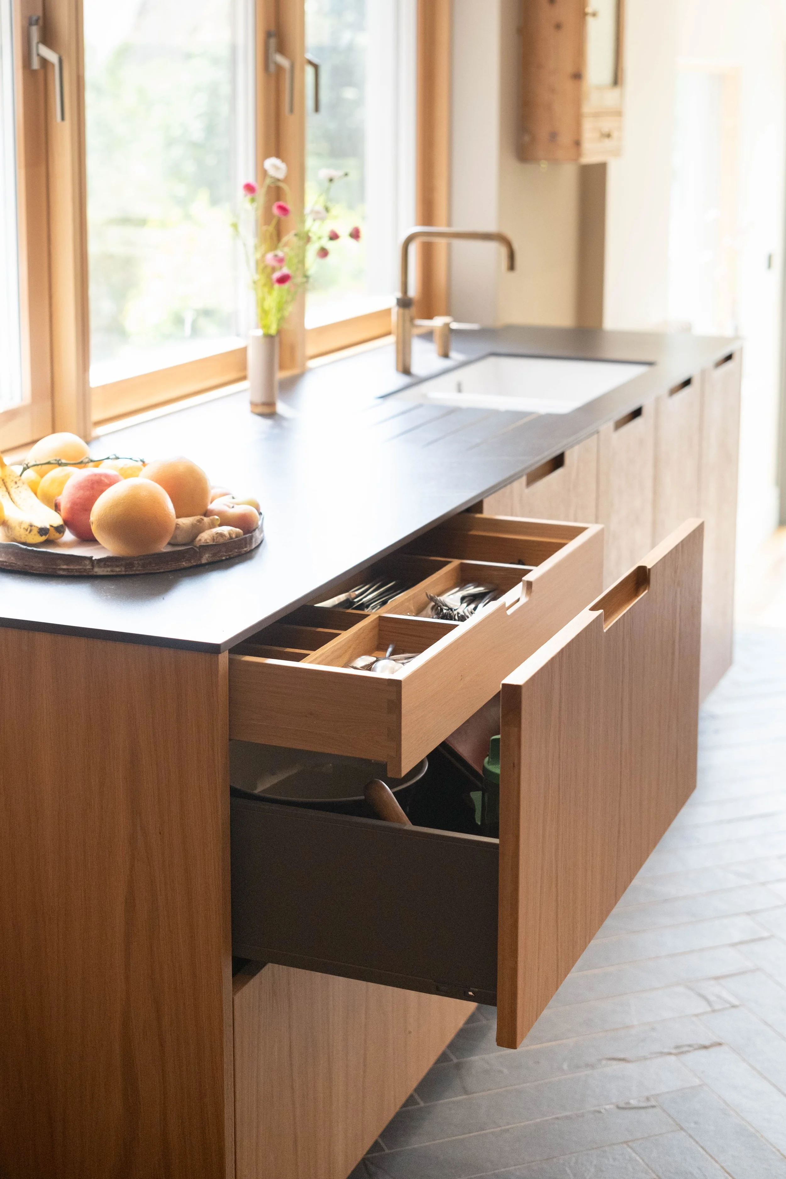 Open kitchen drawer with silverware, a wooden tray of fresh fruits including apples, bananas, and peaches, a window with natural light, a vase with pink and white flowers, a bathroom faucet, and wooden cabinets.