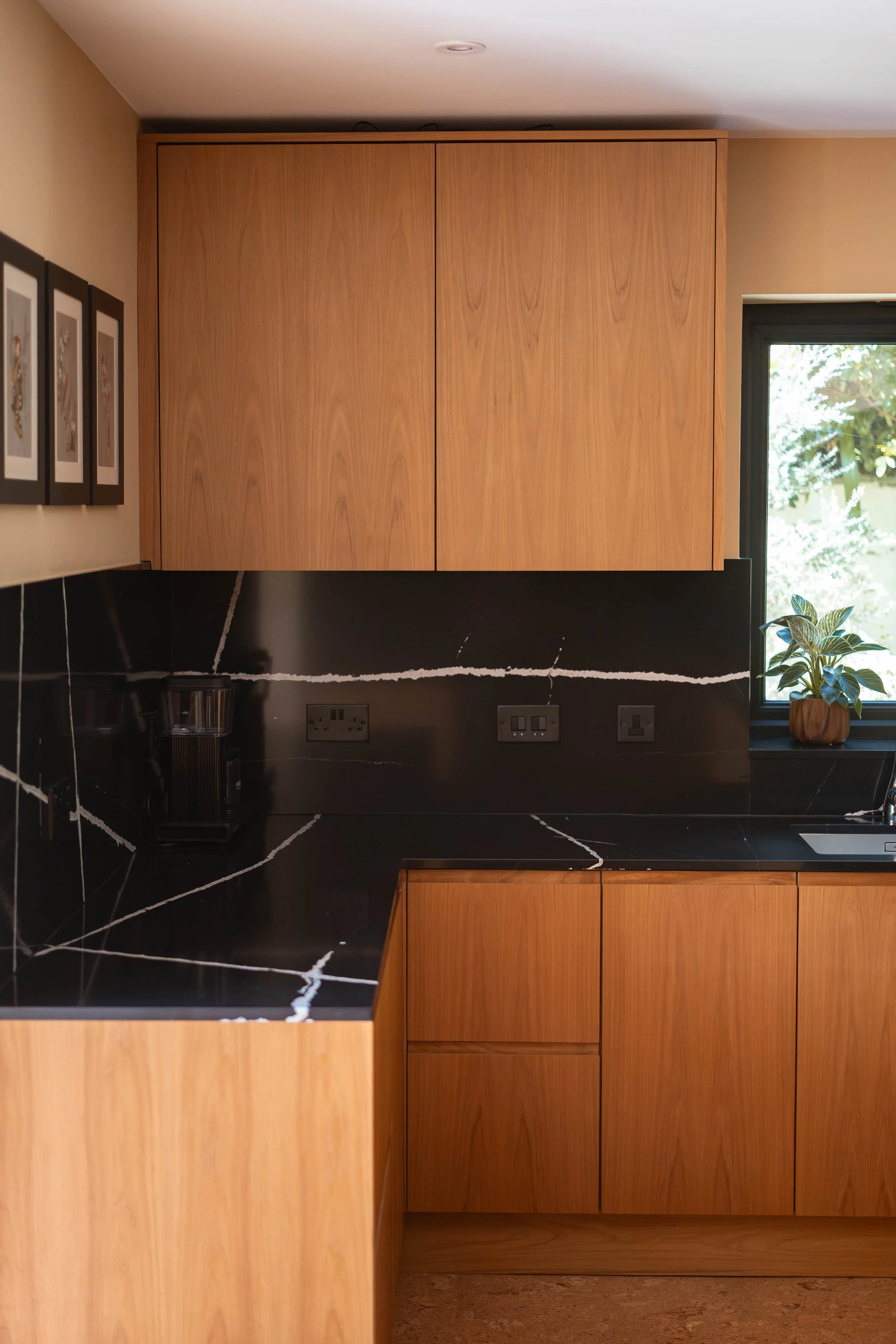 A kitchen with wooden cabinets, black marble countertops with white veining, and a window with greenery outside. There are three framed pictures on the beige wall to the left.