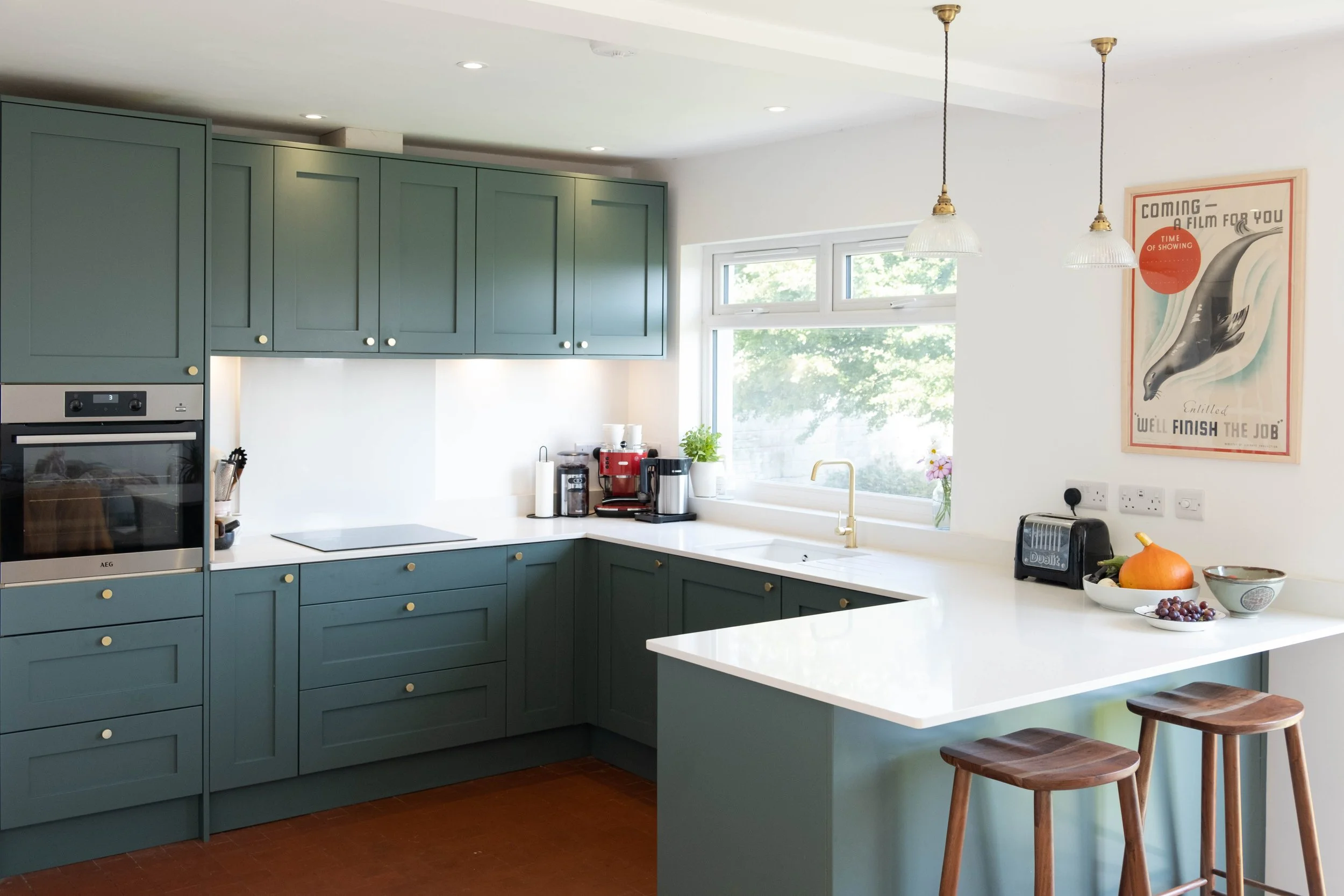 Modern kitchen with green cabinets, white countertops, a double oven, and a window with a view of greenery. There are hanging pendant lights, a poster on the wall, and a bowl of fruit on the counter.