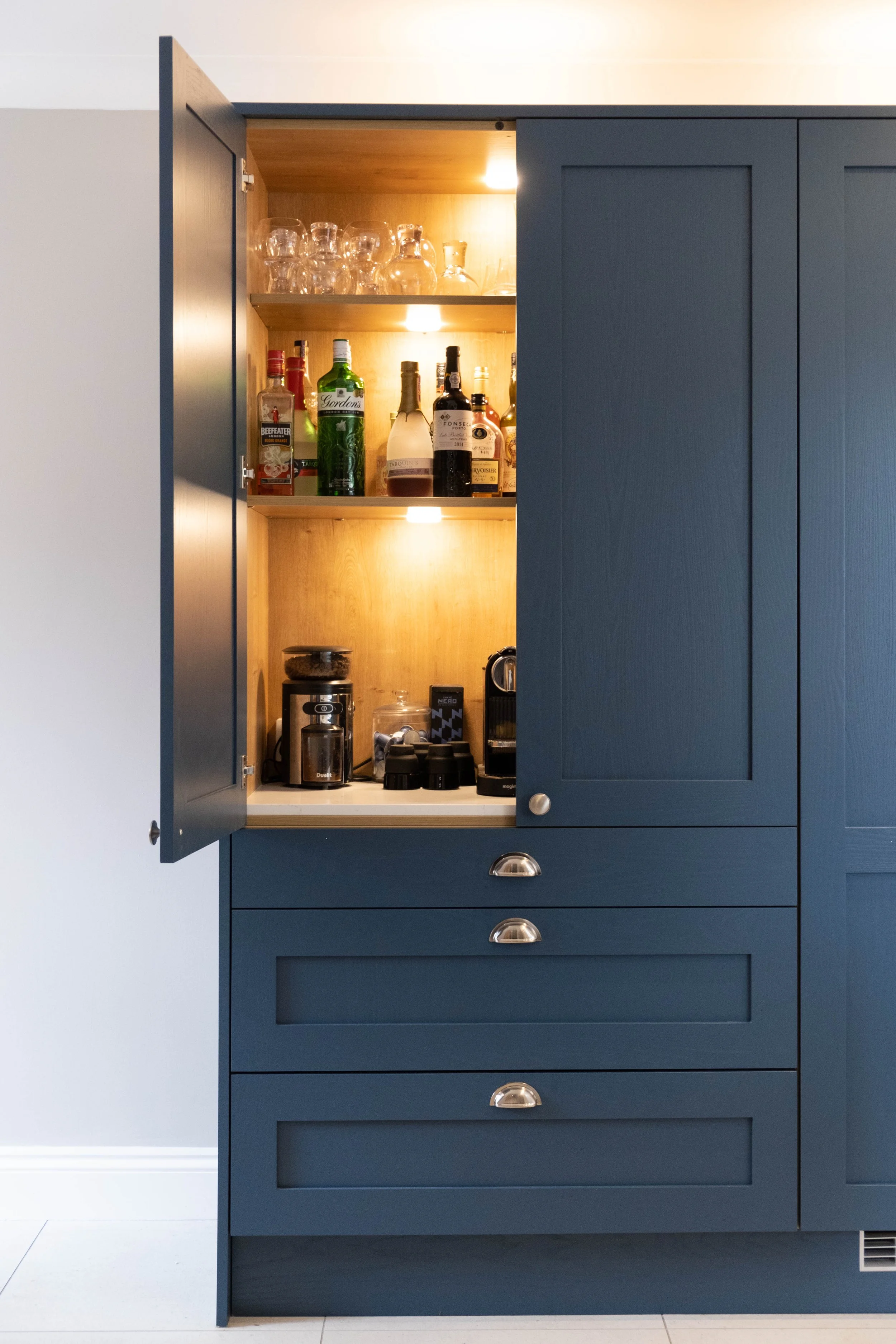 Open blue kitchen cabinet with three shelves containing glassware, liquor bottles, and coffee-making appliances.