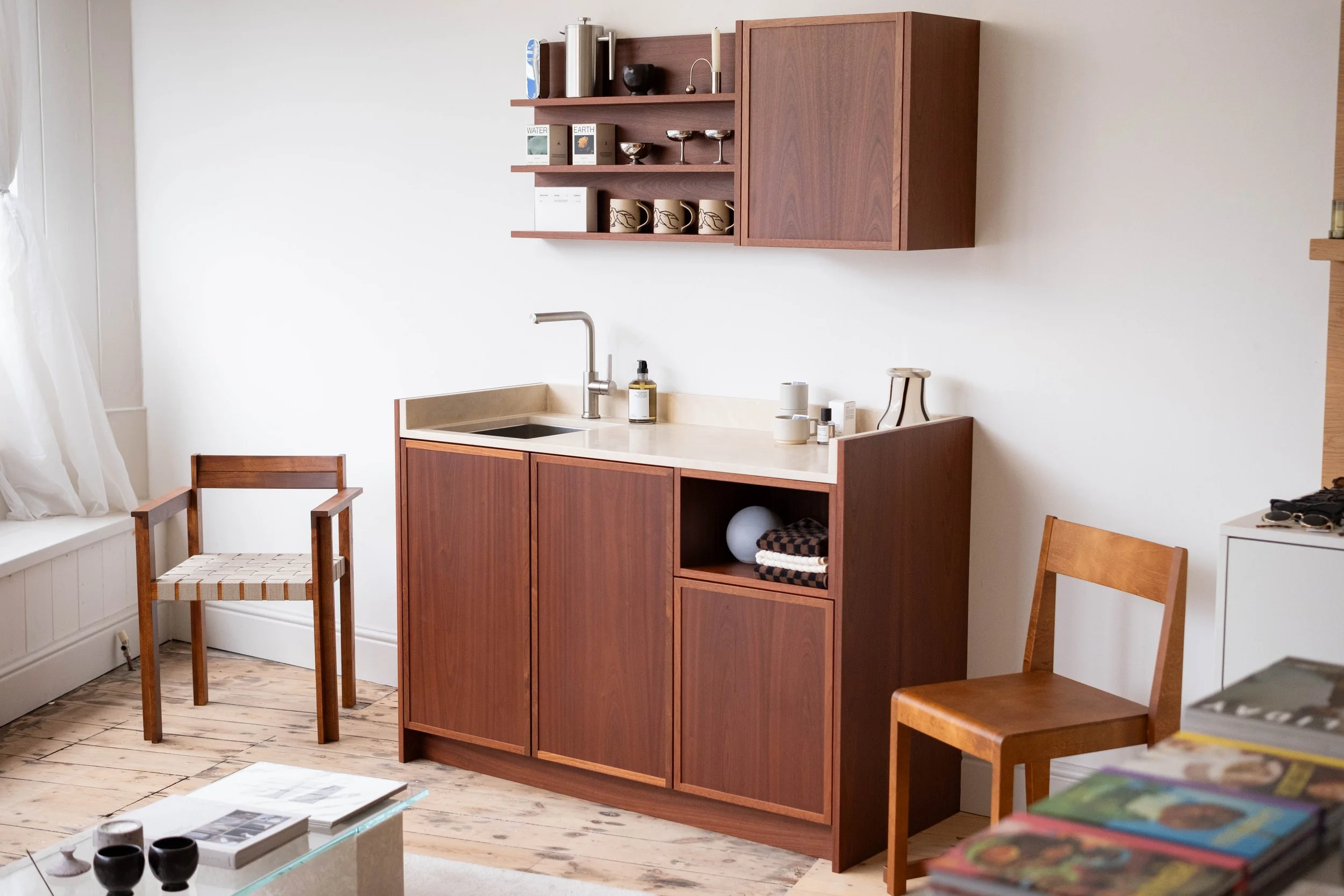 A cozy kitchen corner featuring a wooden cabinet with open shelves, holding cups, bowls, and decorative items, above a cream-colored countertop with a sink and a faucet, surrounded by minimal furniture and light window curtains.