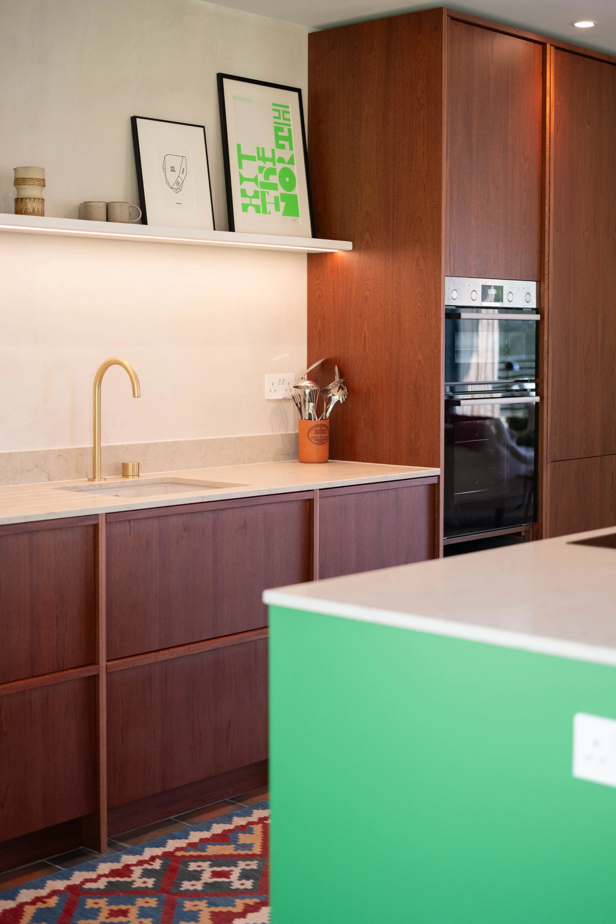 A kitchen with a wooden cabinet, a white counter with a gold faucet, a colorful patterned rug, a green counter in the foreground, and framed artwork on a shelf above the counter.