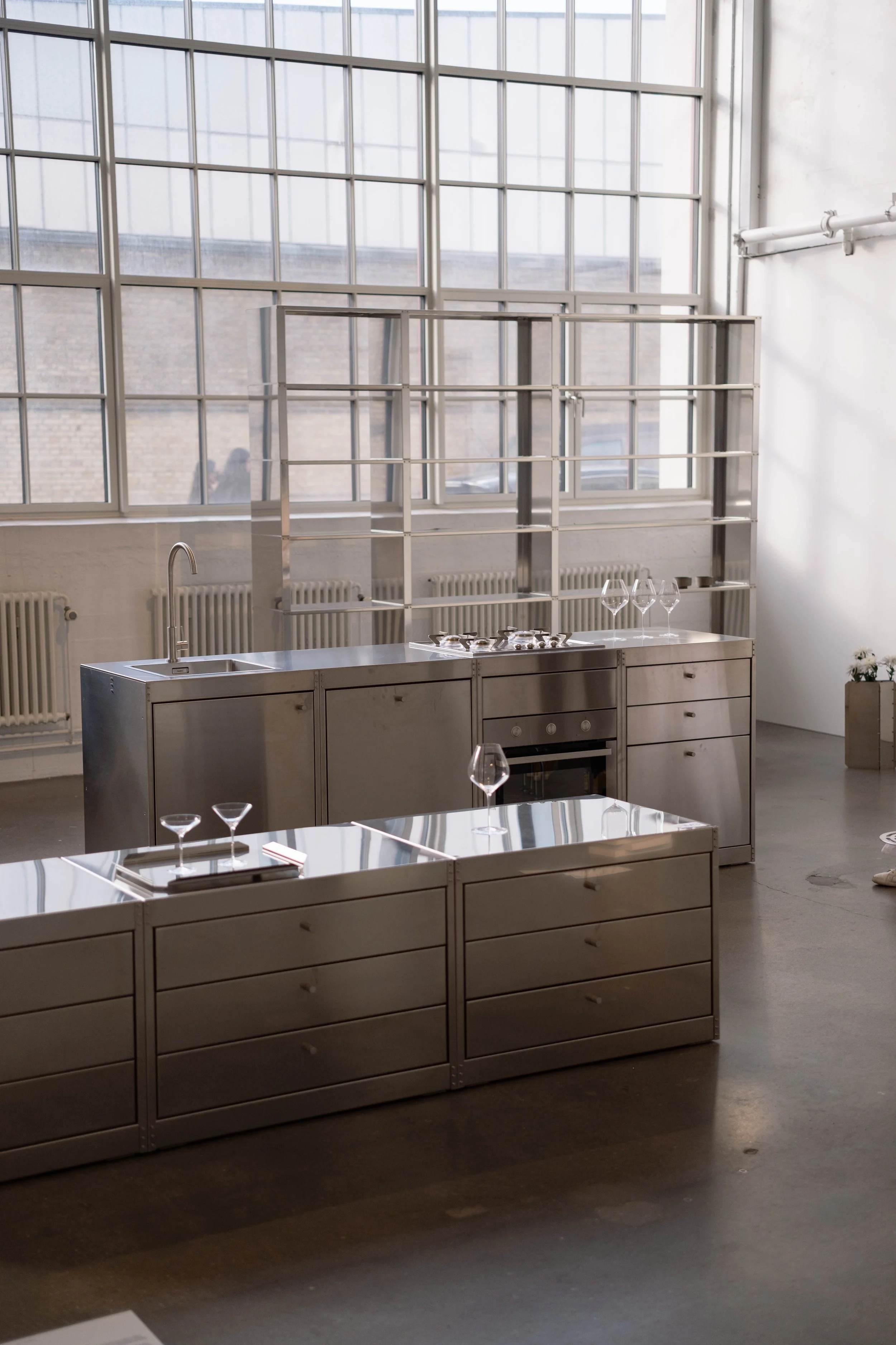 Modern kitchen with metal cabinetry, large window with grid pattern, stainless steel countertops, wine glasses, and an oven.