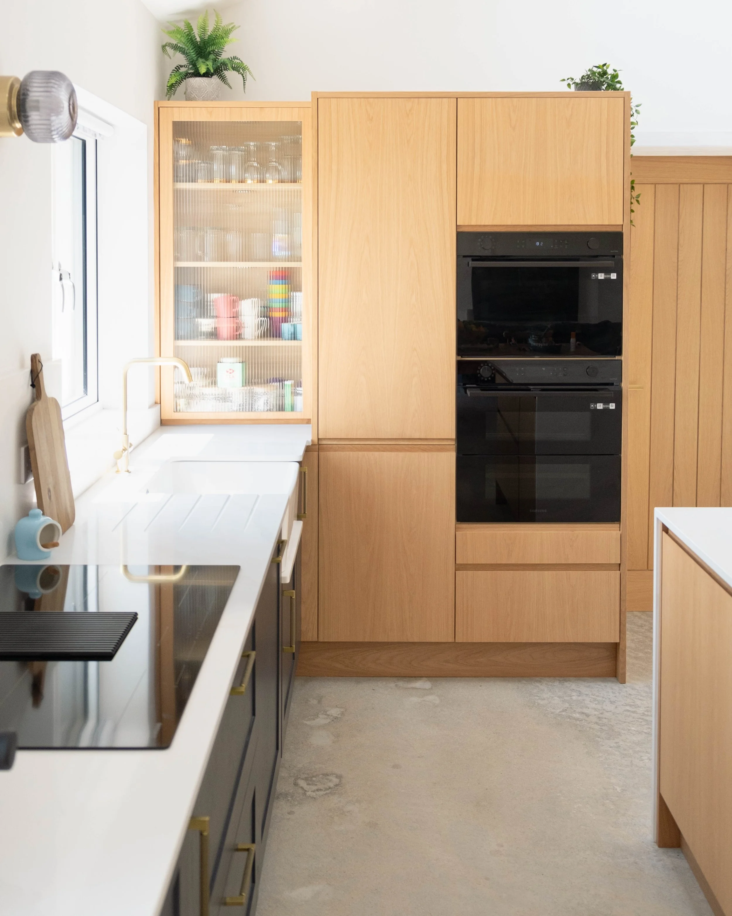 View of a modern kitchen with wooden cabinets, a built-in black oven, a white countertop, a window with a white frame, potted plants on top of the cabinets, and a textured, light-colored floor.