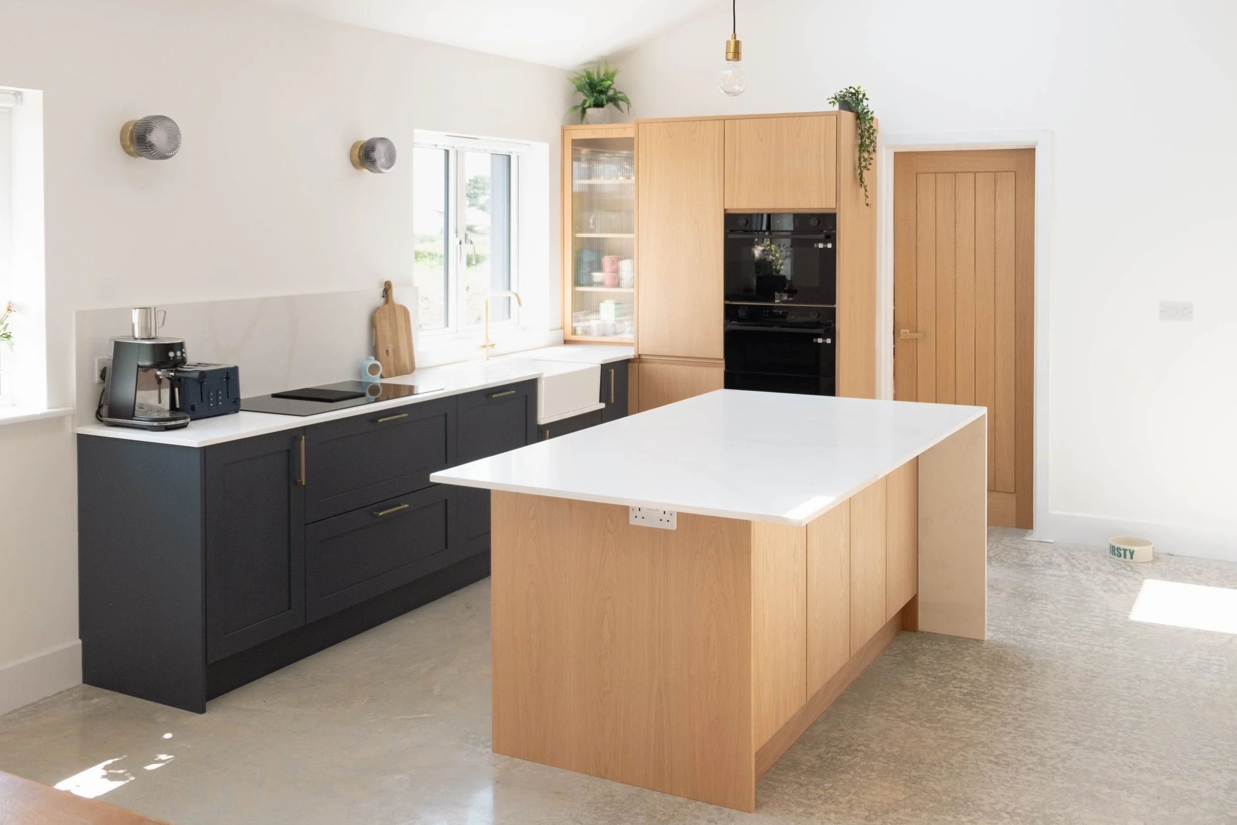 A modern kitchen with black lower cabinets, light wood upper cabinets, a white countertop, and a kitchen island made of wood. There are two windows, a potted plant on top of a tall cabinet, and a hanging light bulb.