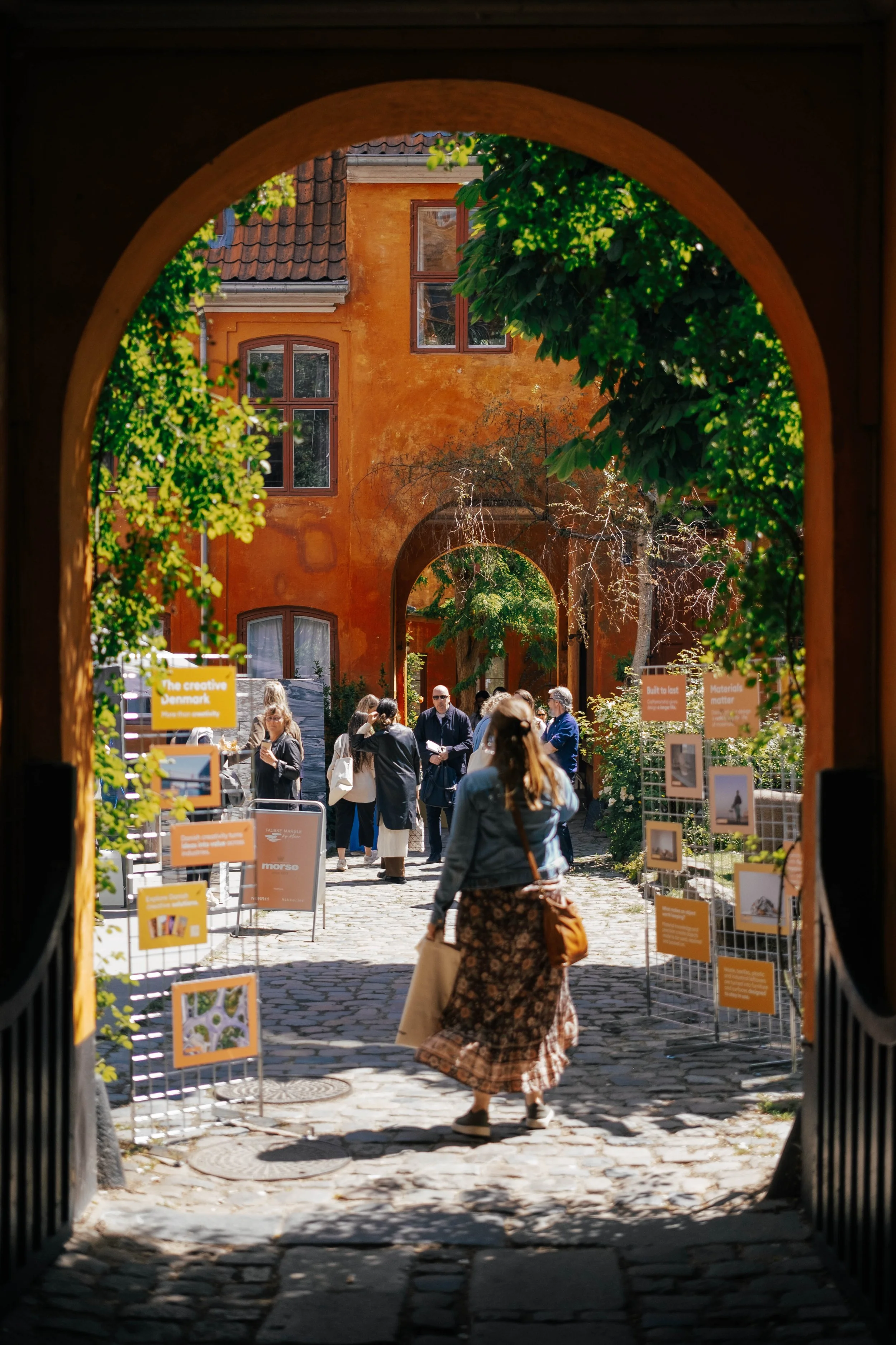 View of a cobblestone street through an arched opening with people walking and informational display signs on both sides, leading to a yellow-orange building with large windows and a tiled roof, surrounded by green trees.