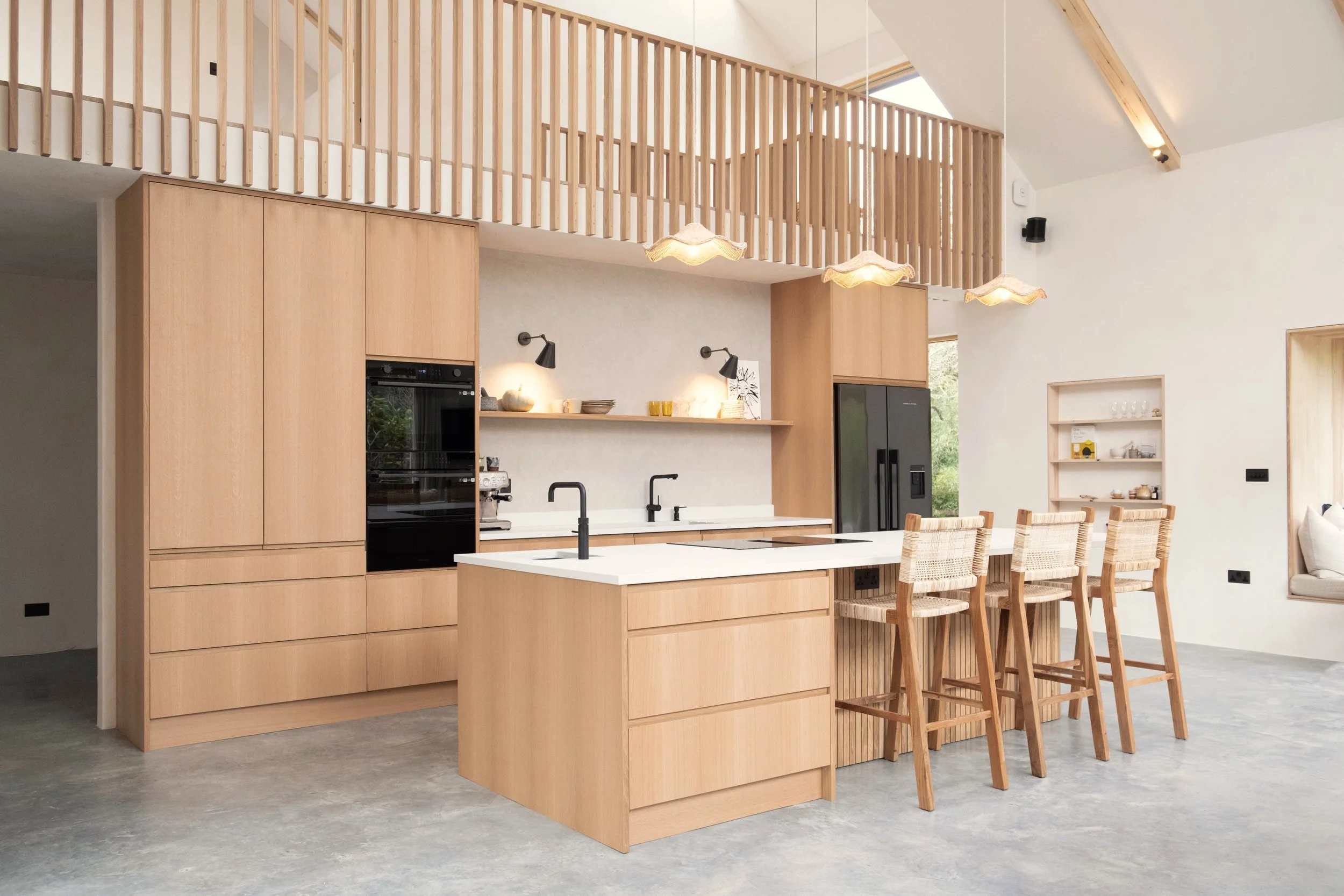 Open-concept kitchen and dining area with light wood cabinetry, a white island with black fixtures, a row of wooden chairs at a dining table, and a loft area with wooden railing.