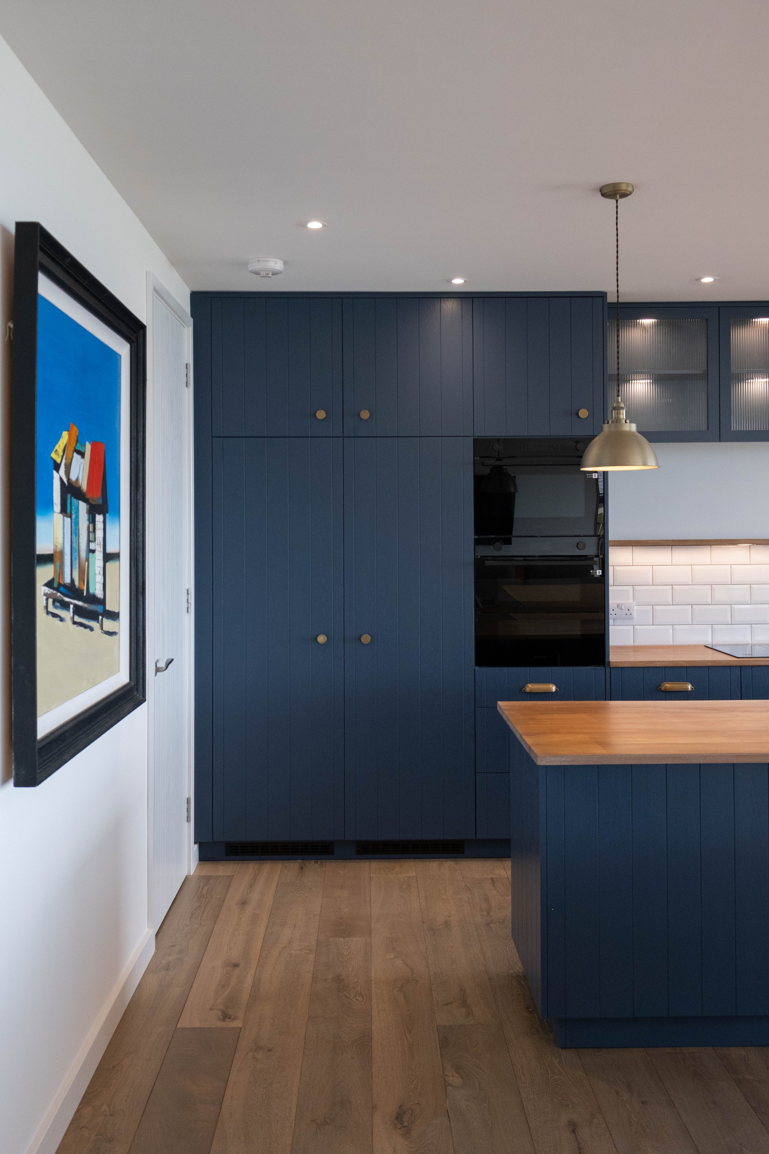 A modern kitchen with navy blue cabinets, a built-in oven, white subway tile backsplash, and a wooden countertop. A hanging pendant light is above the counter, and a colorful framed artwork is on the adjacent white wall.