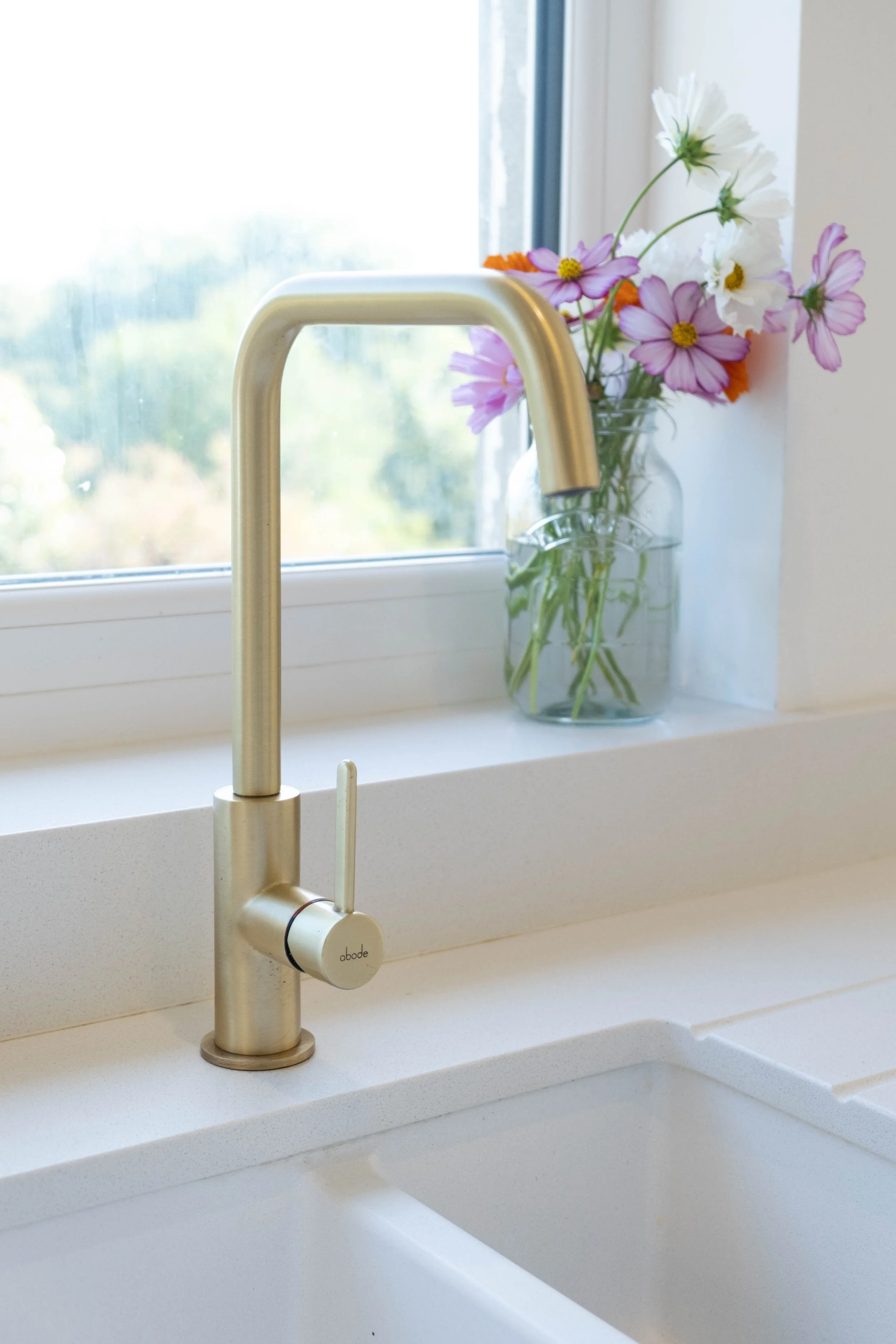 A modern kitchen faucet installed on a white sink with a vase of colorful flowers on the windowsill in the background.