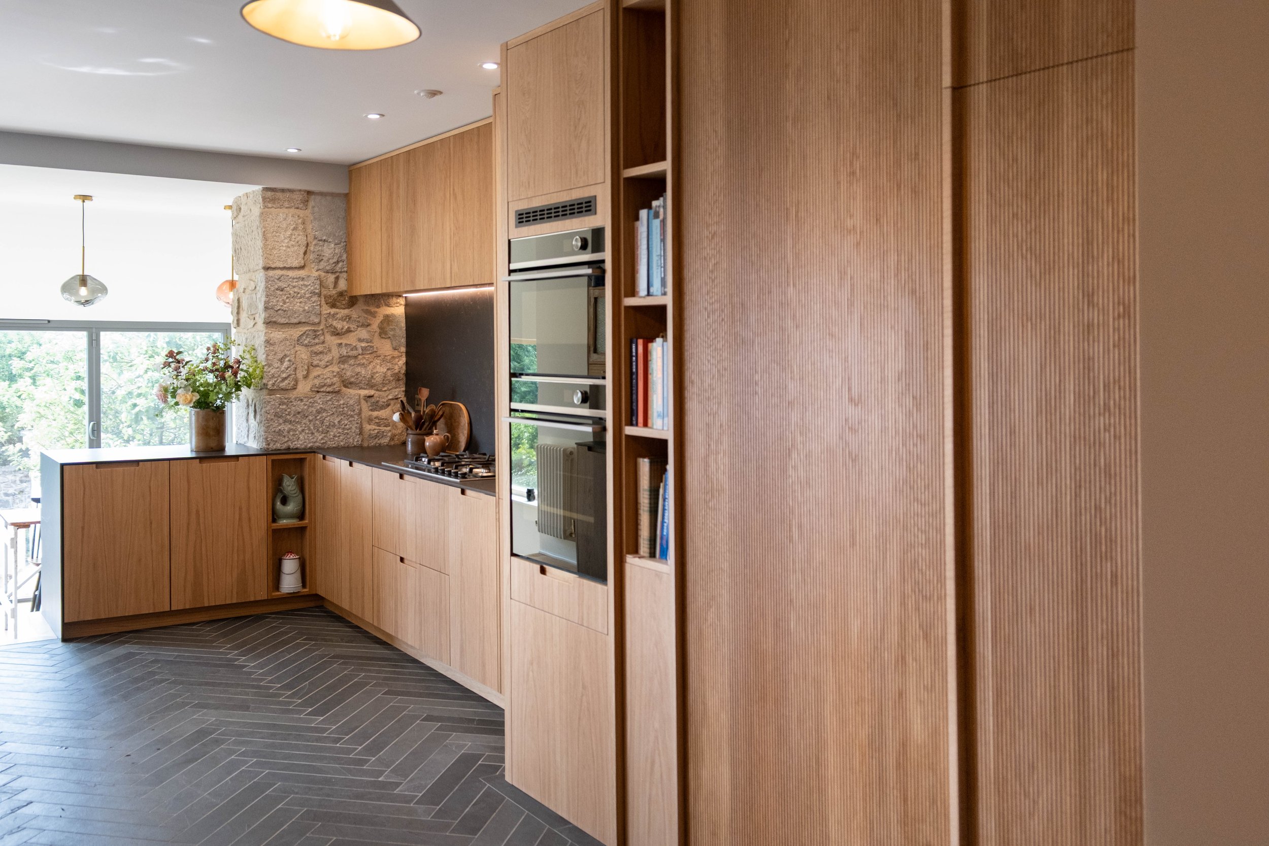Modern kitchen with wooden cabinetry, stone wall feature, glass doors, and decorative pendant lights, with a vase of flowers on the counter.