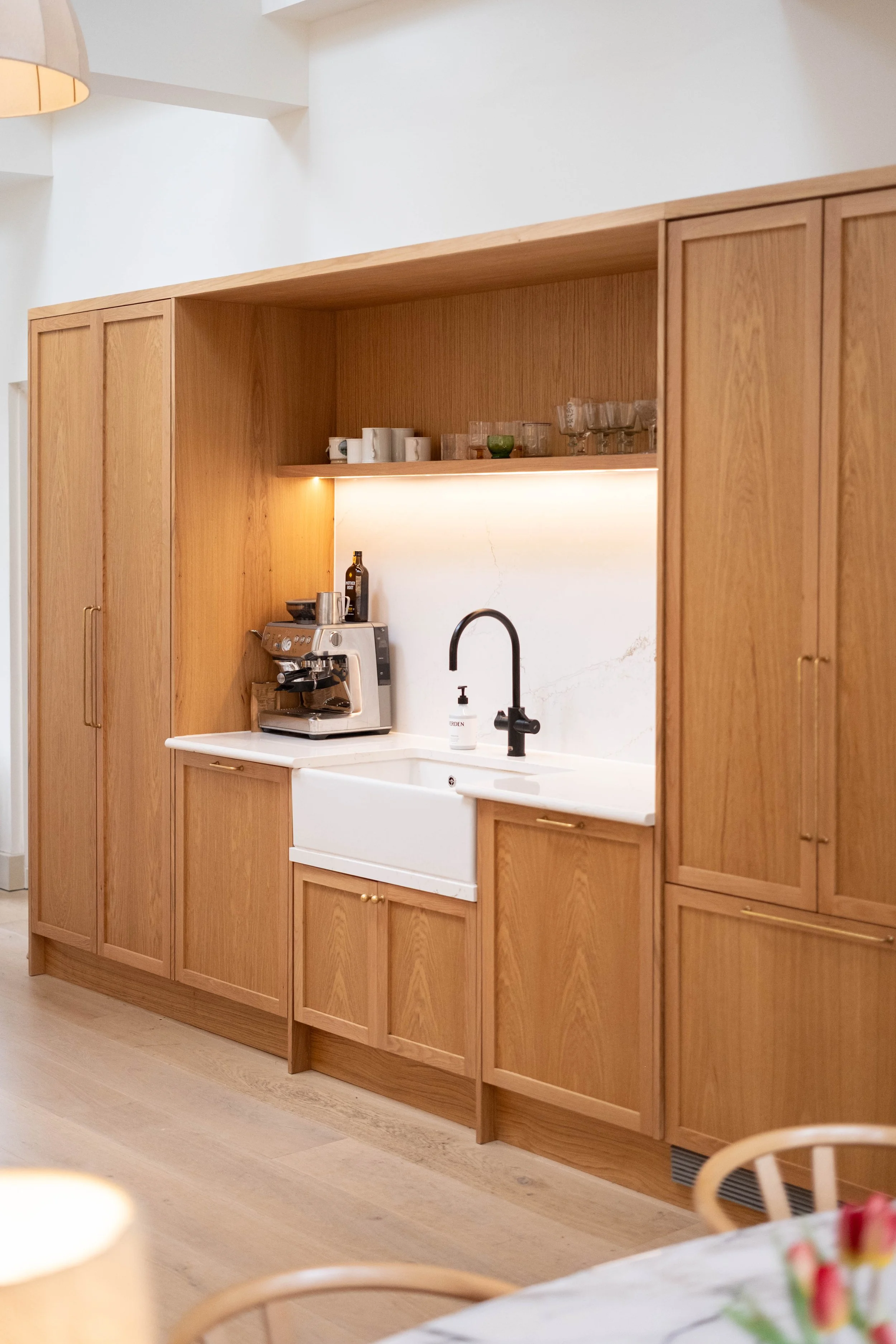 A kitchen with wooden cabinetry, a white farmhouse sink, and a coffee machine on the counter. There are cups and glasses on a shelf above the counter.