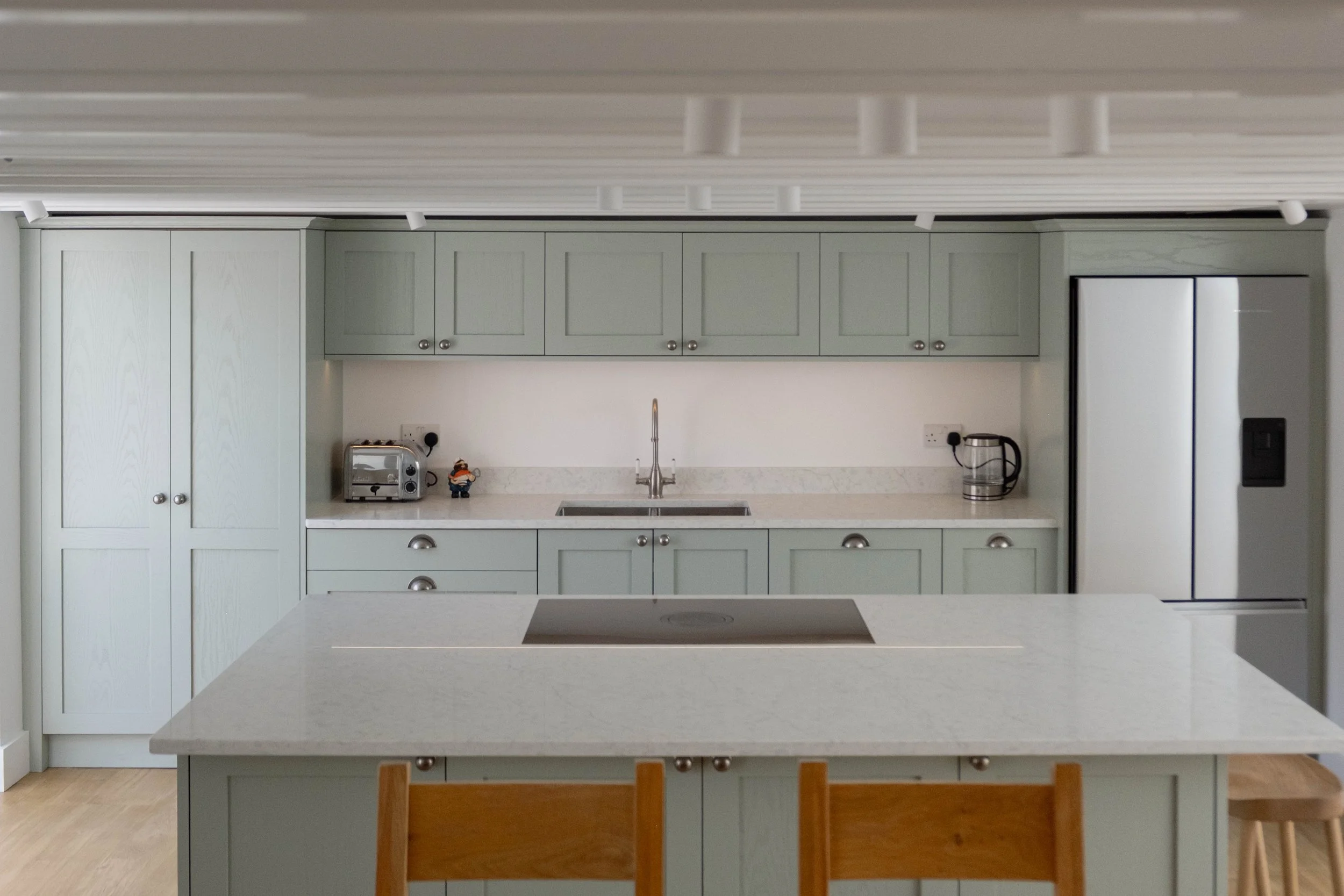 Modern kitchen with pale green cabinets, white countertops, a kitchen island, and a stainless steel refrigerator, with small appliances on the counter.