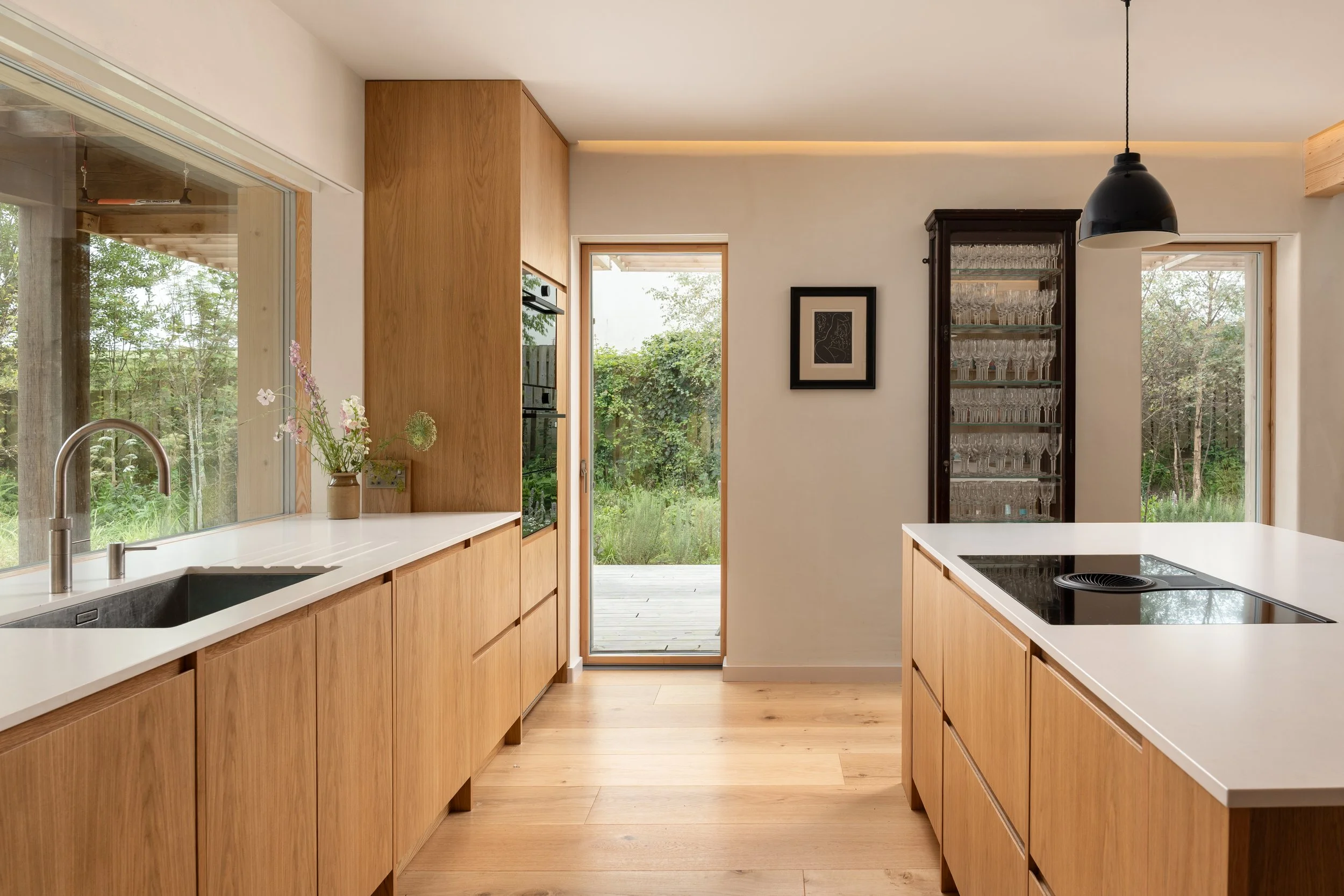 Modern kitchen with wooden cabinets, white countertops, a black stovetop, glass cabinet with glassware, a framed artwork on the wall, and large windows showing a green outdoor landscape.