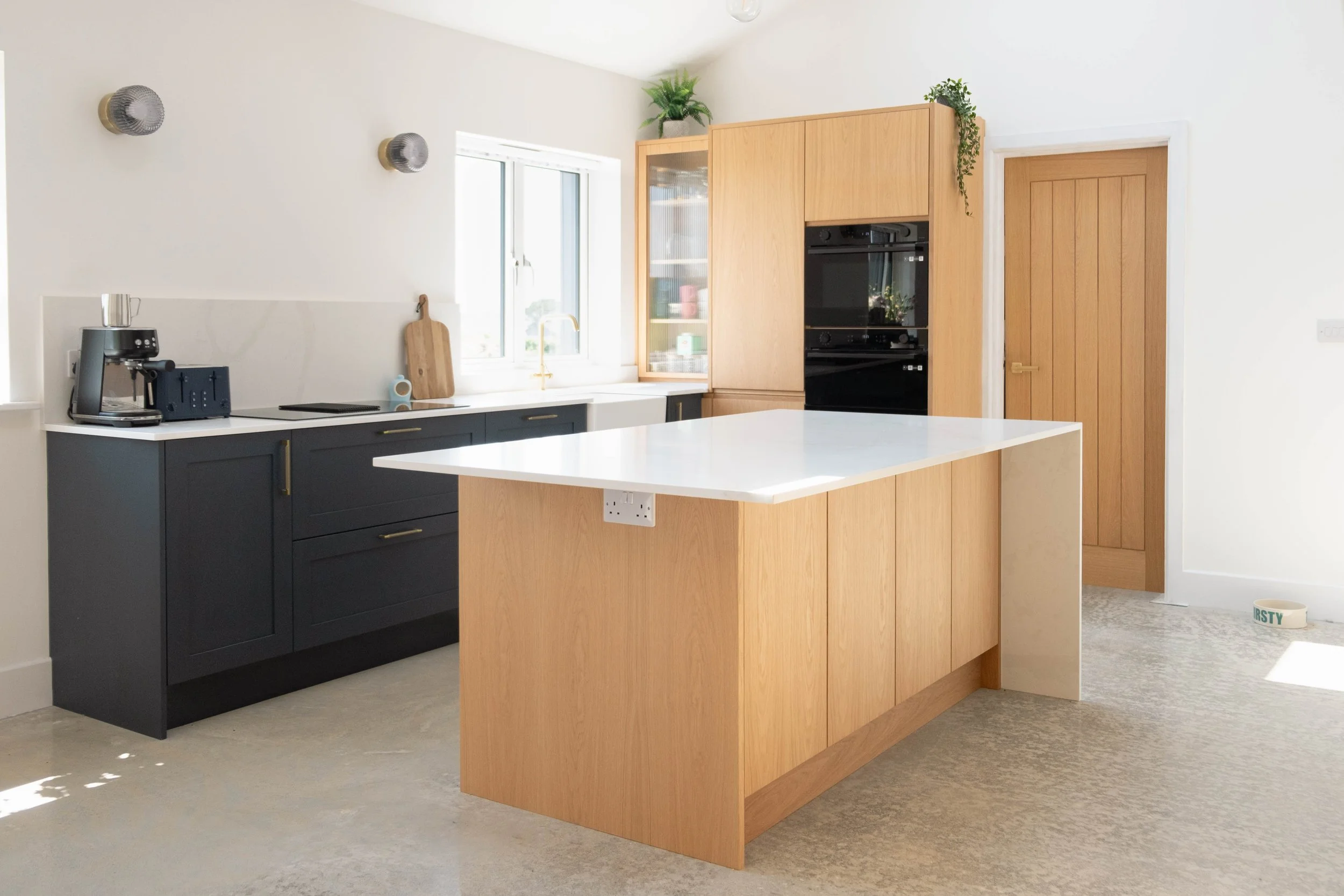 Modern kitchen with dark blue lower cabinets, a white countertop, and wooden upper cabinets. There is a window above the sink, and a coffee machine on the counter. A wooden cutting board hangs on the wall next to the window, and there are some plants